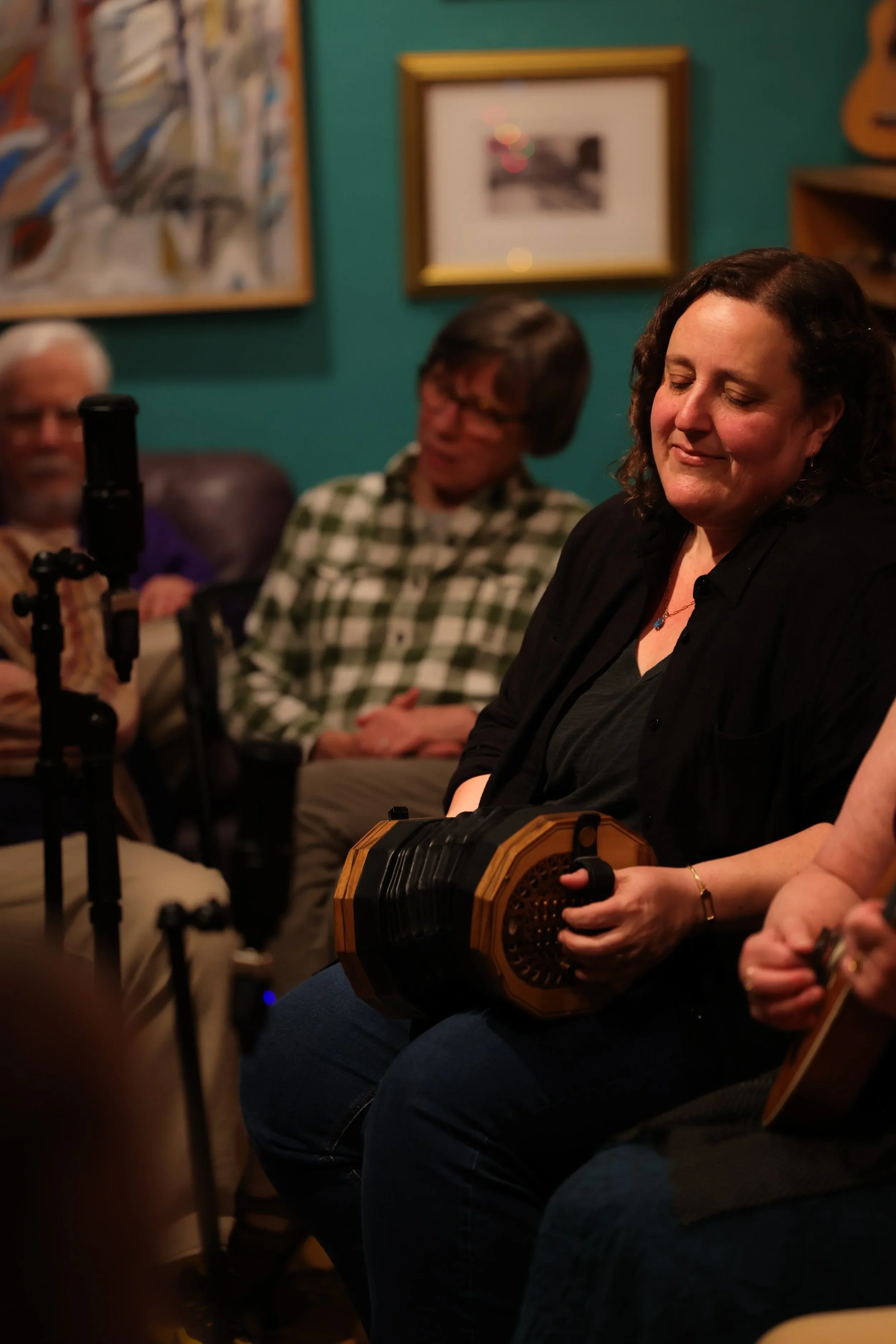 A woman with dark, curly hair playing a concertina during a gathering in a cozy room with teal walls. Other people are sitting nearby, with framed artwork hanging on the wall behind.
