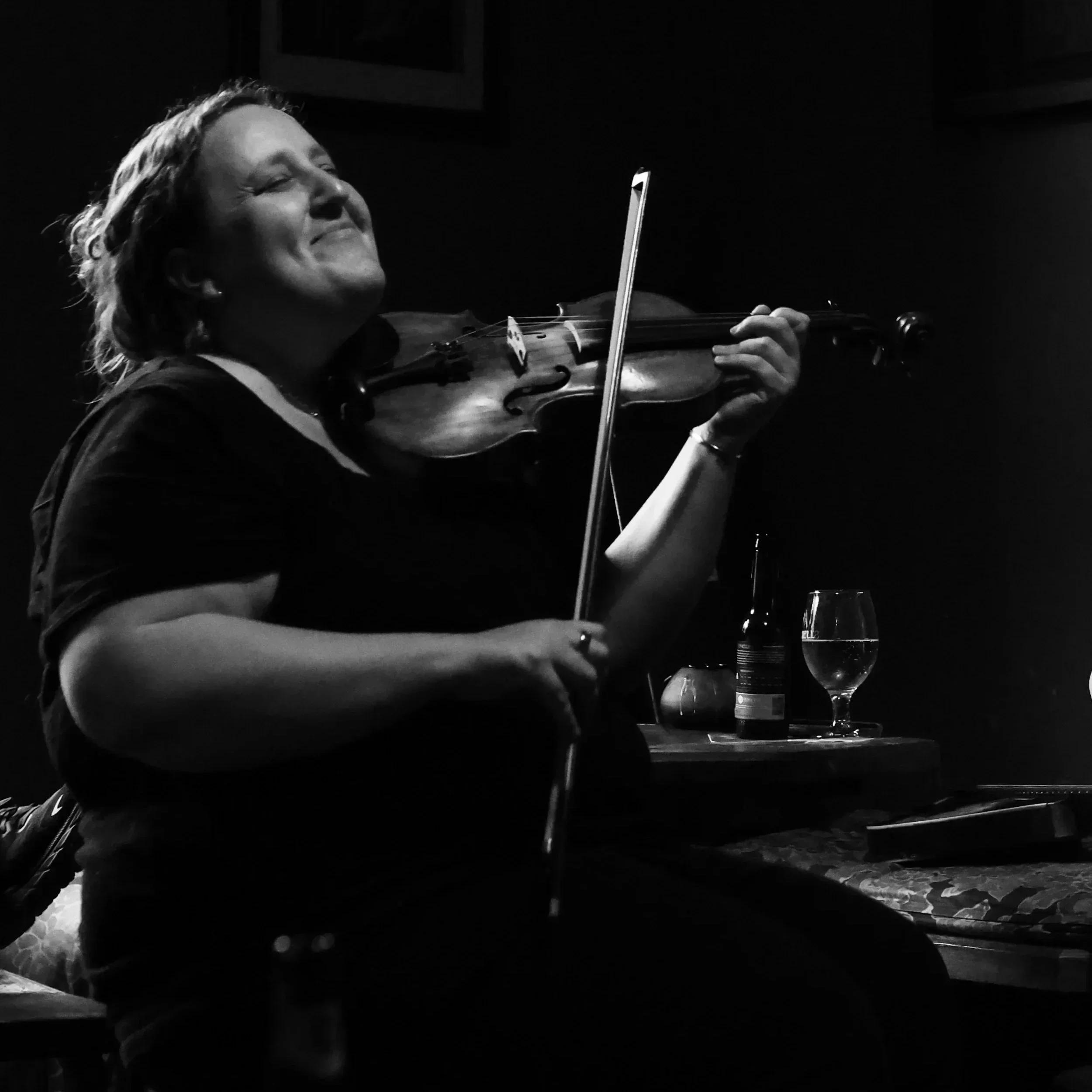 Black and white photo of a woman passionately playing the violin, with her eyes closed and a content expression, seated at a table with a glass of water and a bottle.