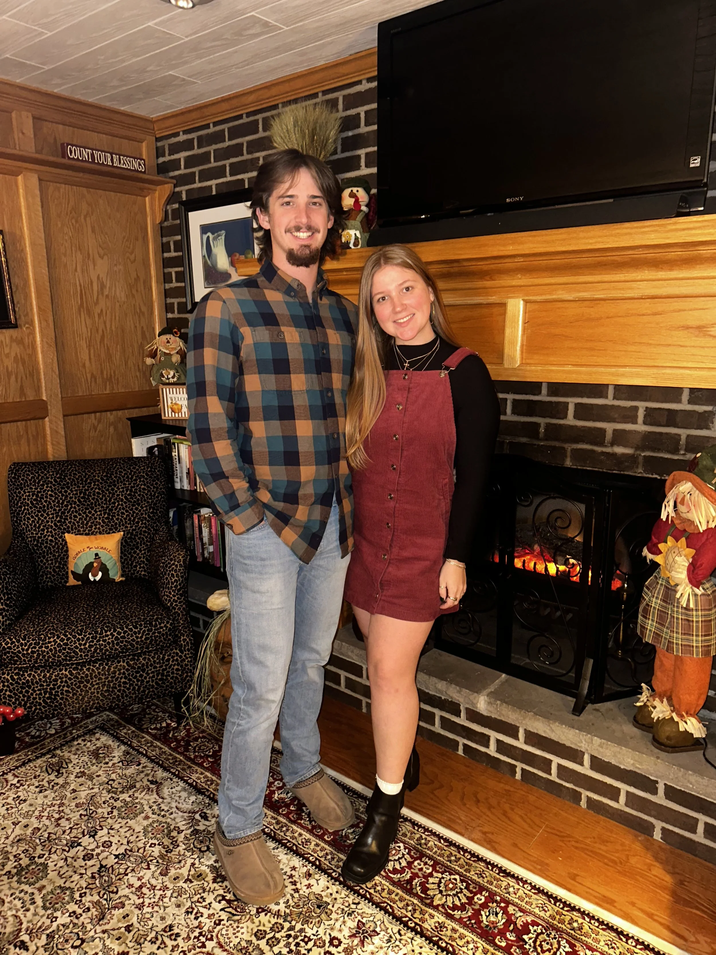 A young man with dark hair and a goatee, wearing a multicolored plaid shirt and light jeans, standing next to a young woman with long red hair, wearing a black long-sleeve top and a red corduroy dress, in a cozy living room with a fireplace and holiday decorations.