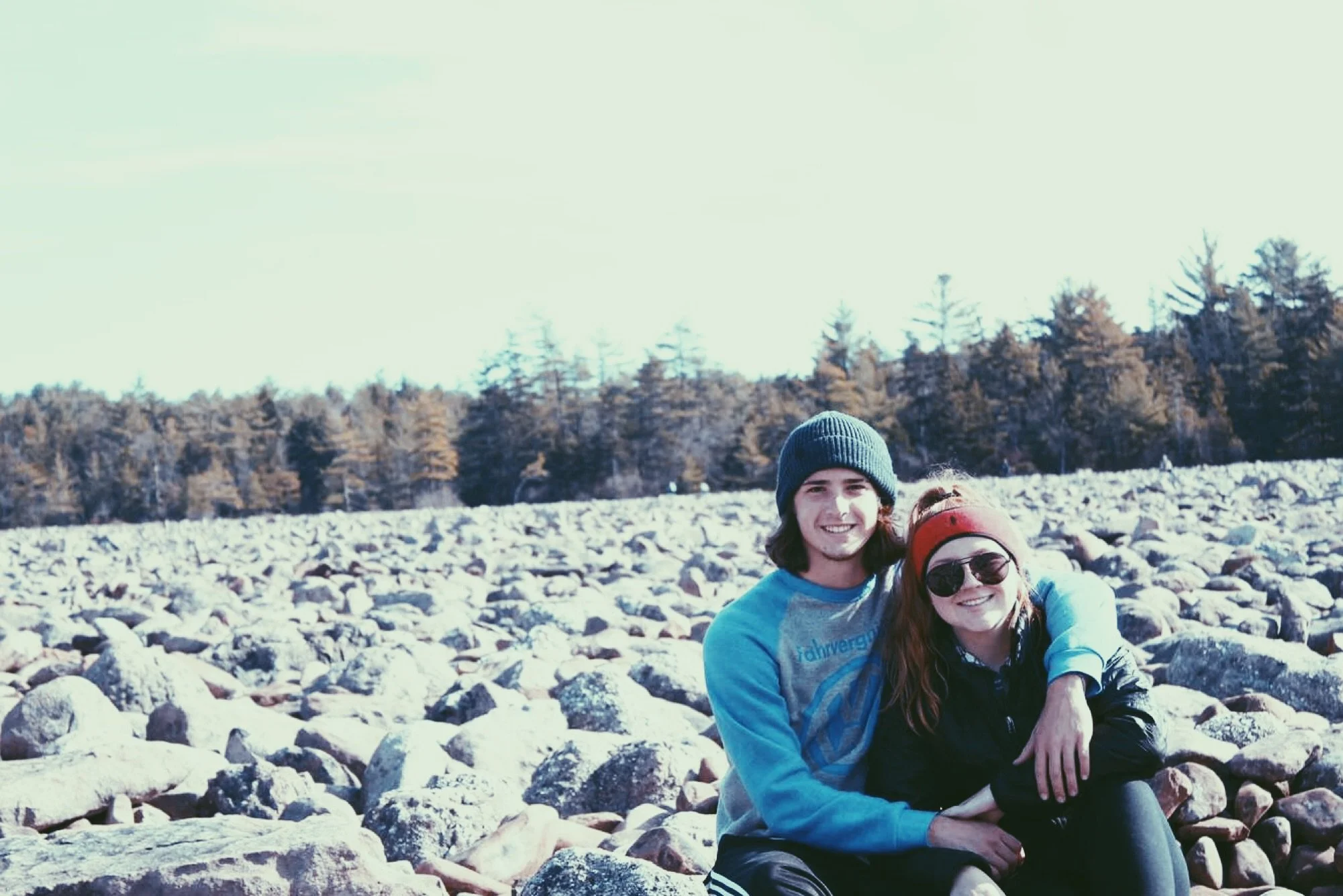 A young man and woman smiling and sitting on rocks in an outdoor setting with trees in the background, wearing casual clothing and beanies.
