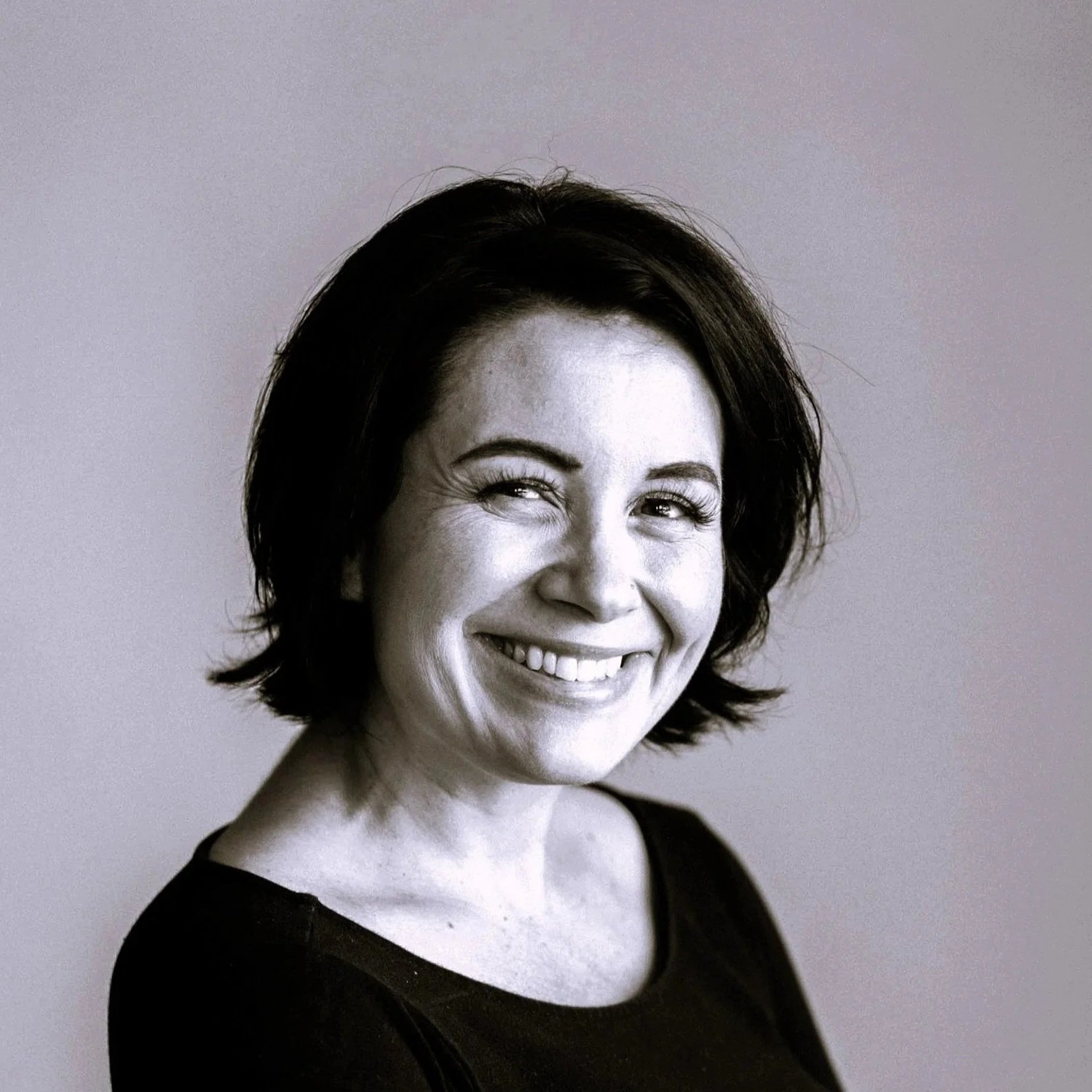 Black and white portrait of a woman with short, dark hair, smiling with her teeth showing, wearing a black top in front of a neutral background.