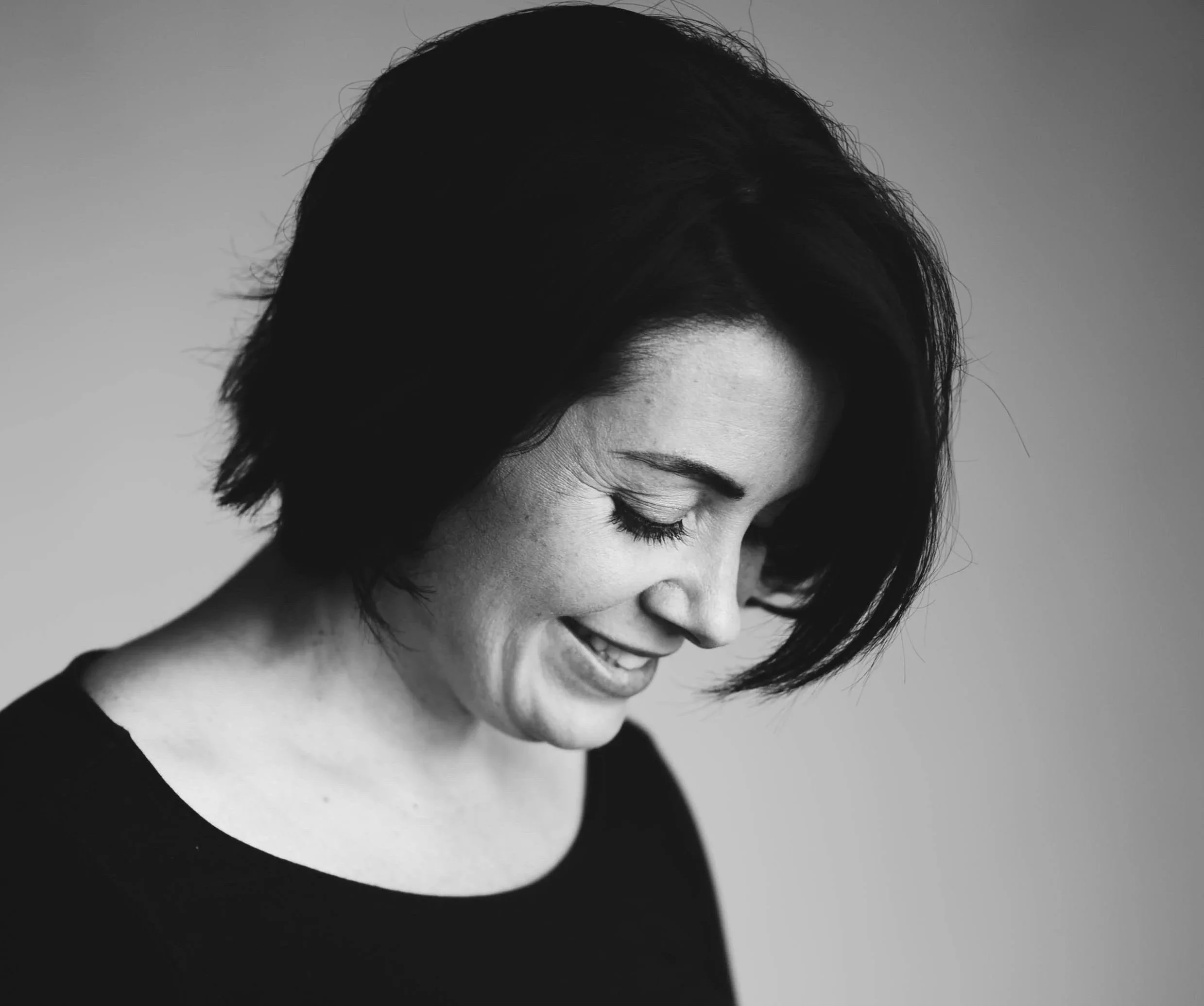 Black and white portrait of a woman with short dark hair, smiling with her eyes closed, wearing a black top, against a neutral background.