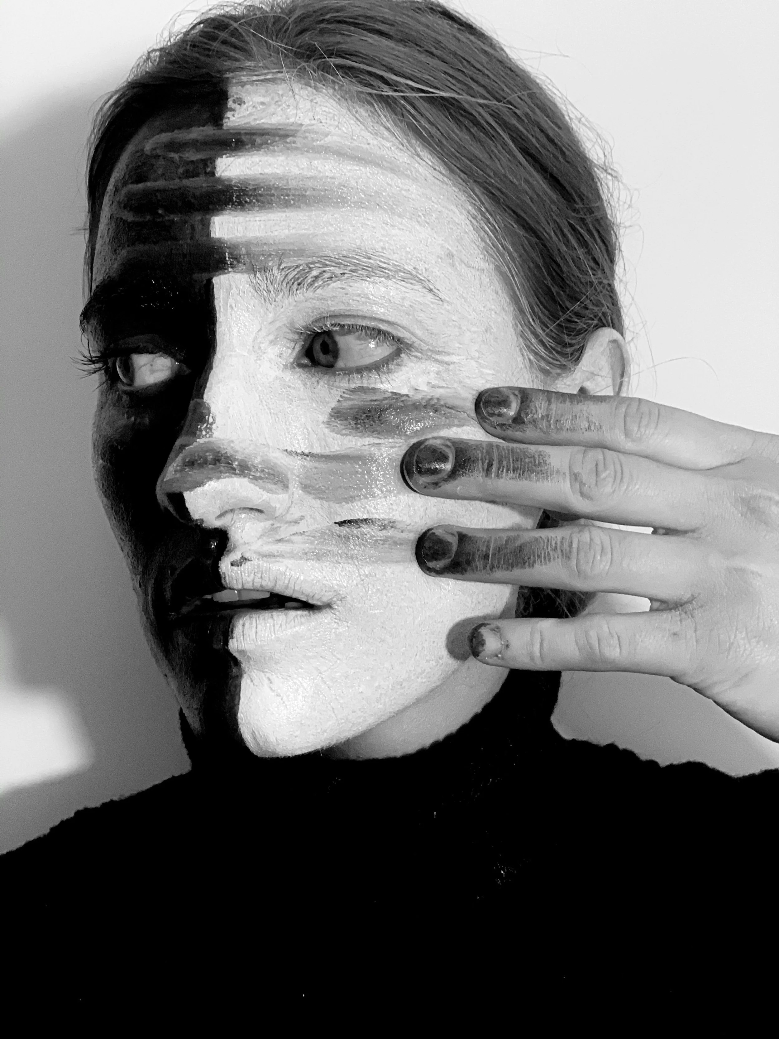 A black and white photo of a woman with makeup. Her face is partially covered by her hand, with fingers painted in dark nail polish. She has an intense gaze, and her hair is tied back. The makeup appears smudged or painted on.