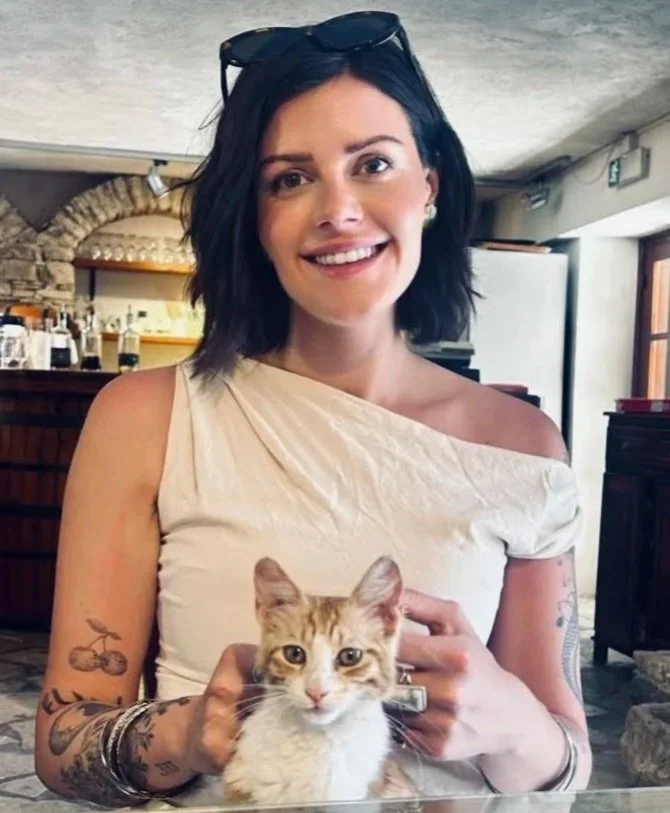 Woman with dark hair, wearing a light-colored off-shoulder top, sitting in a rustic indoor space, holding a young ginger and white cat