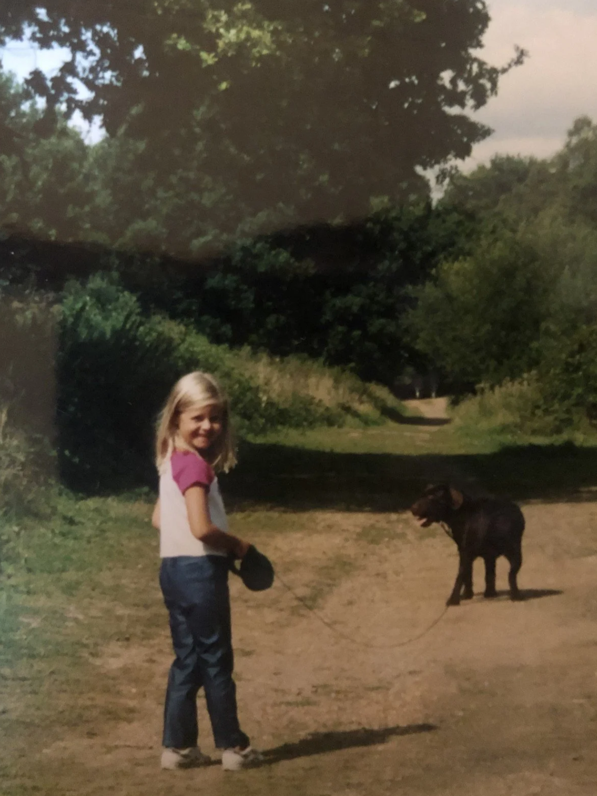 A young girl with blonde hair, wearing a white and pink shirt, blue jeans, and white shoes, standing outdoors with a black dog on a leash in a natural setting with trees and a dirt path.