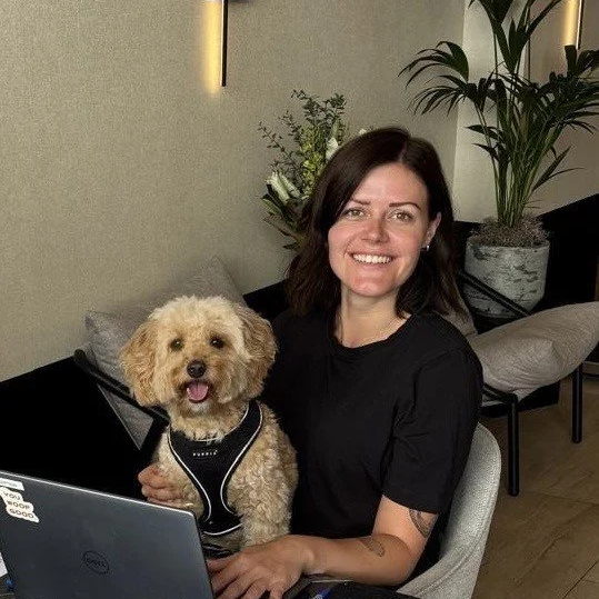 A woman with dark hair smiling and sitting at a desk with a poodle wearing a vest, in a modern indoor setting with plants.