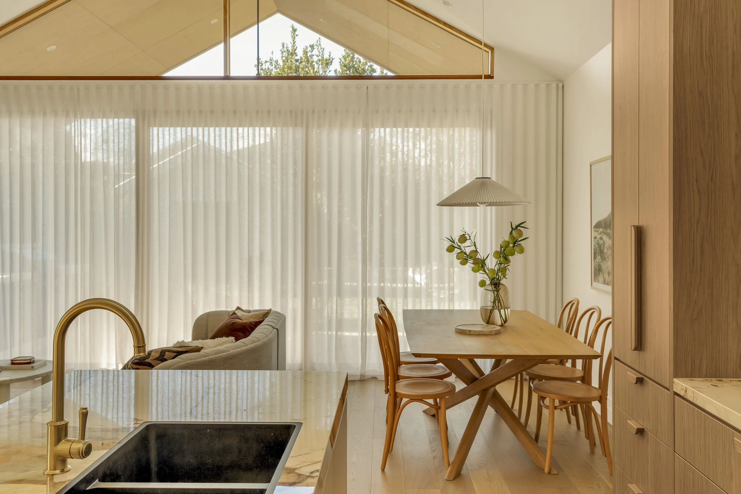 A modern living room with floor-to-ceiling curtains, a wooden dining table with chairs, a vase with flowers, and a elevated ceiling with skylight and trees visible outside.