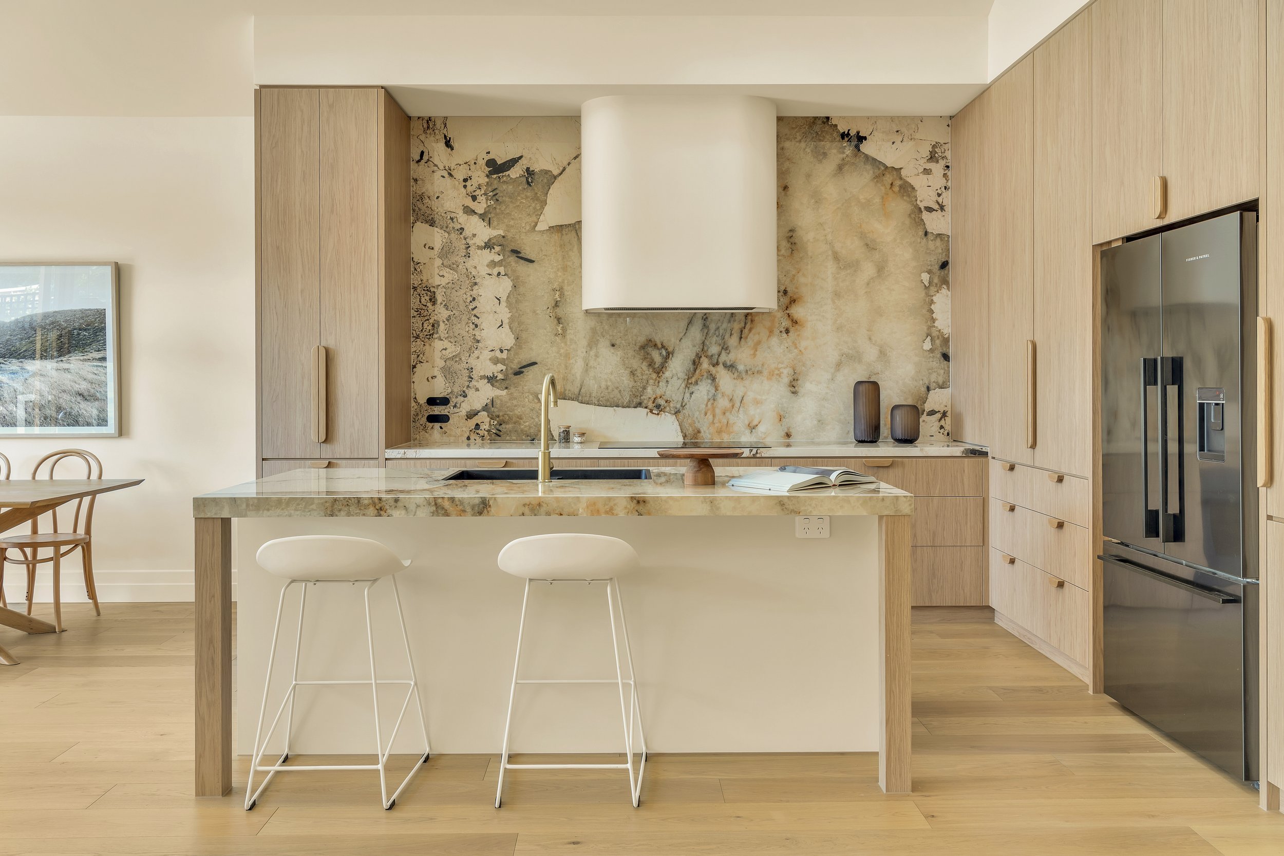 Modern kitchen with light wood cabinets, marble countertop, and a marble backsplash with abstract pattern. Two white bar stools are at the island, and there are decorative vases on the countertop. A doorway on the right reveals a stainless steel refrigerator.