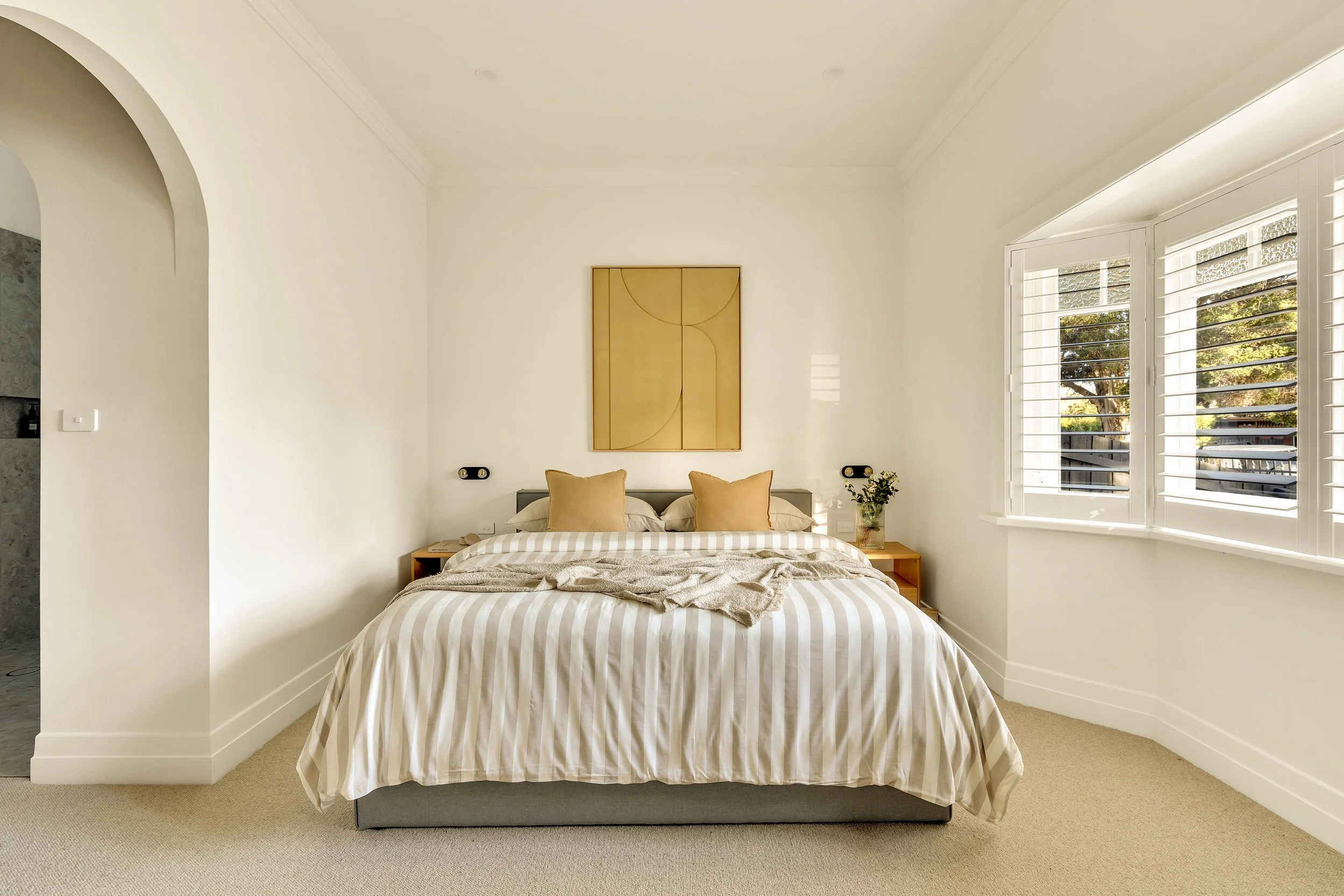 A bedroom with a bed covered in beige and white striped bedding, two beige pillows, and a throw blanket. There's a small wooden nightstand on either side of the bed, decorated with a plant and a book. The wall behind the bed has a wooden abstract art piece, and windows with white blinds are on the right, allowing natural sunlight to illuminate the room.