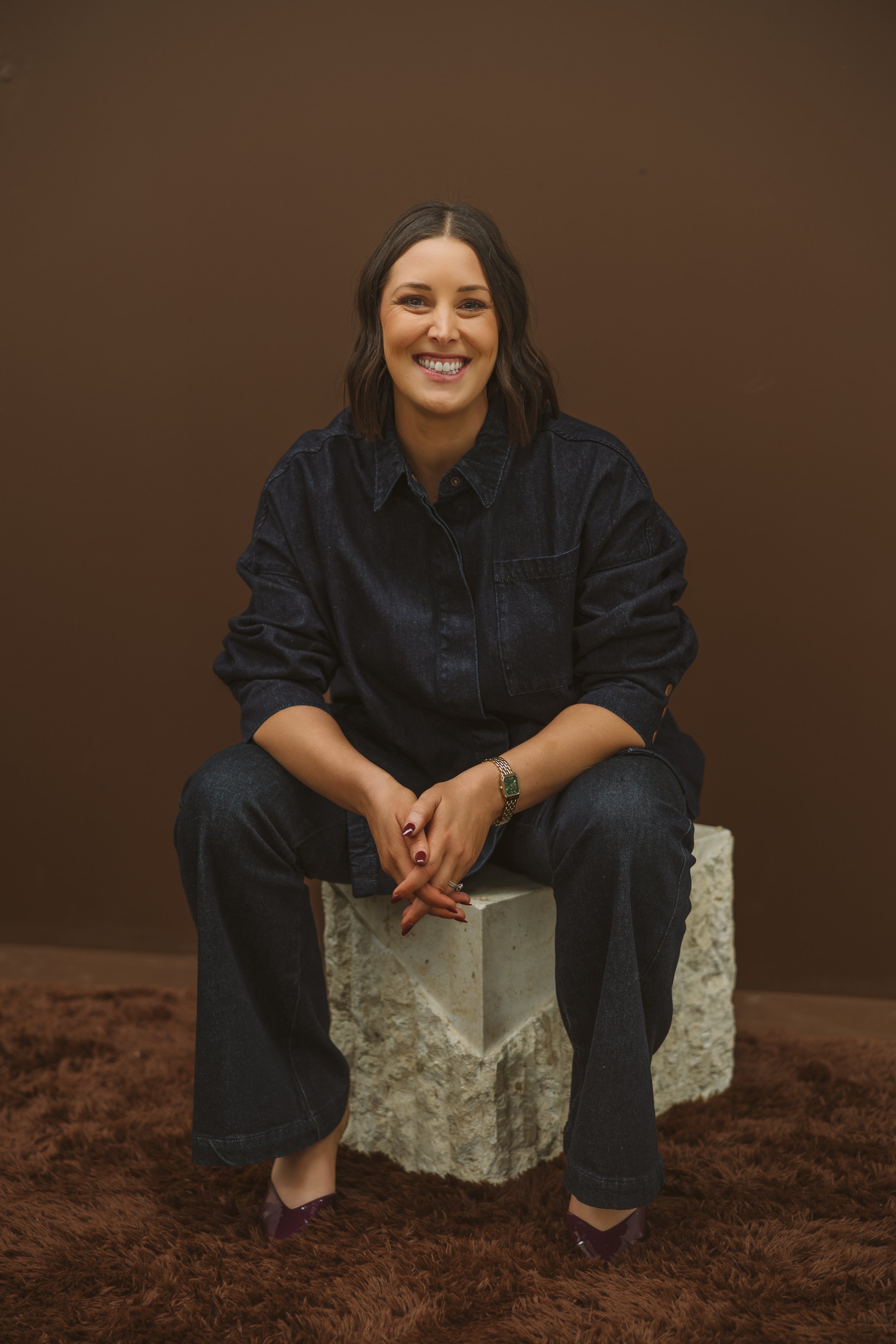 Aimee Bliss of Studio Hue, with shoulder-length dark hair smiling, seated on a white stone block against a brown background, dressed in a black denim jacket, dark jeans, and purple shoes, with a gold bracelet on her left wrist.