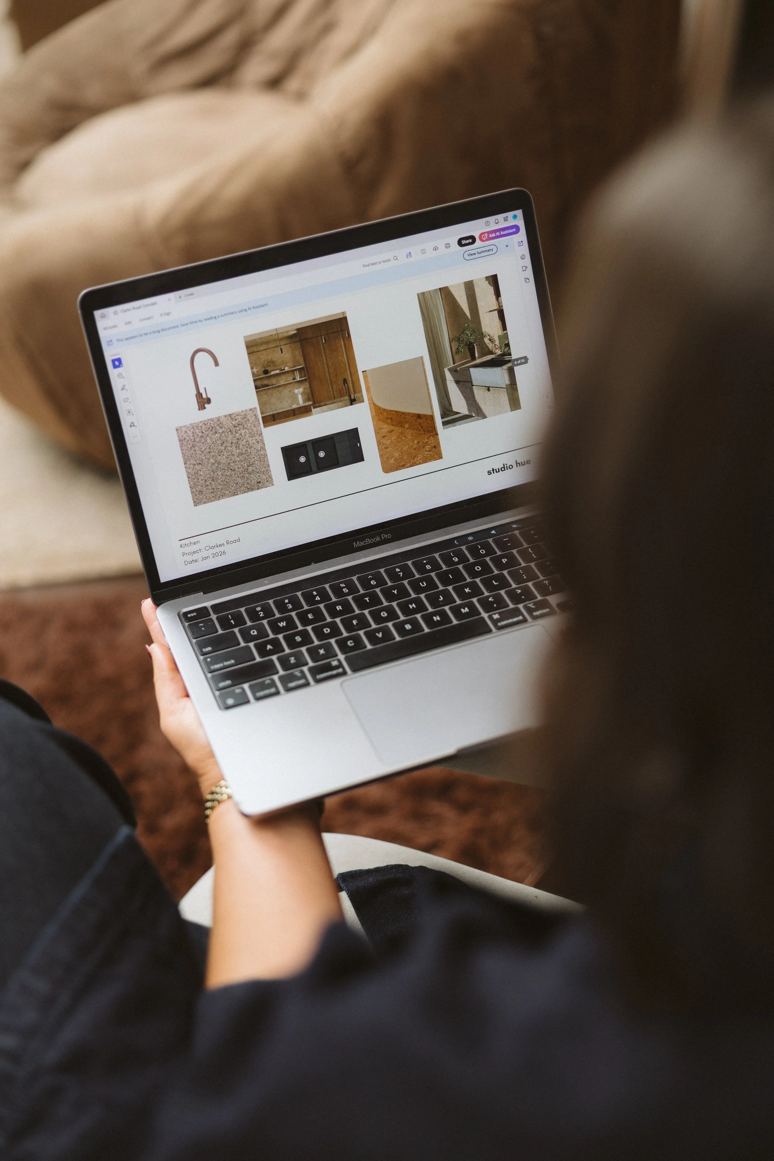 Aimee Bliss of Studio Hue in Gippsland, Victoria, holding a MacBook Pro laptop showing a design project with images of kitchen materials and fixtures. The laptop screen displays color swatches, kitchen cabinetry, countertops, a sink, and plant decor.