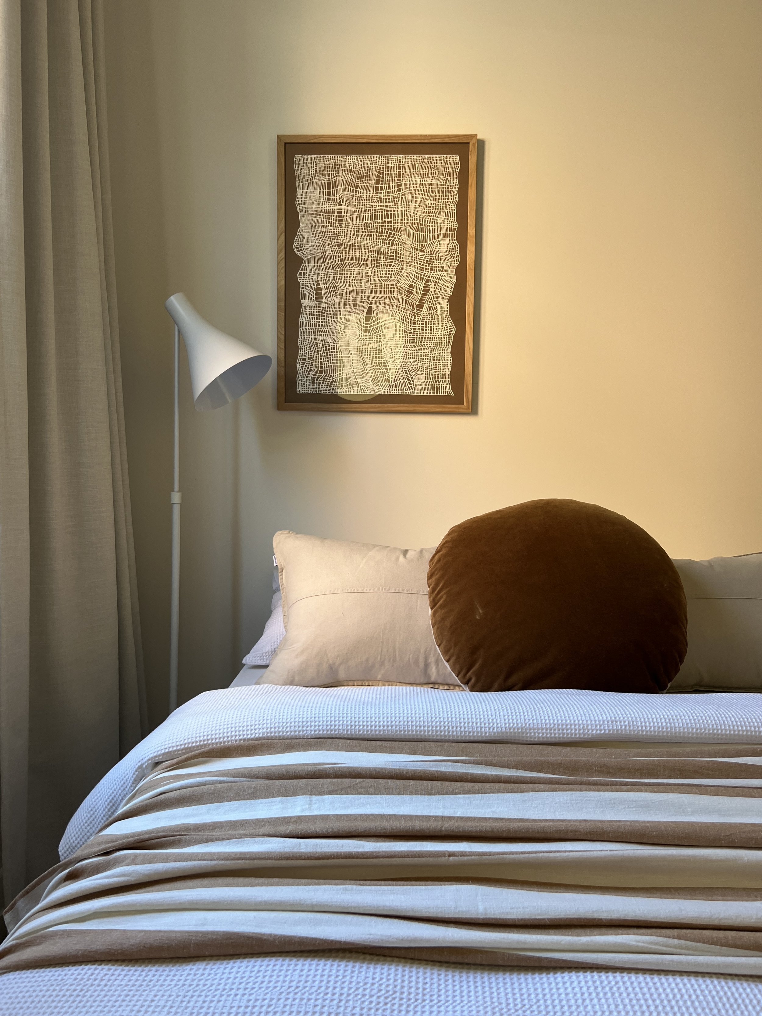 A neatly made bed with white and beige striped sheets, a beige pillow, a large brown cushion, and a headboard with beige pillows. On the wall above is a framed piece of art featuring an intricate white line pattern on a dark background. To the left, 