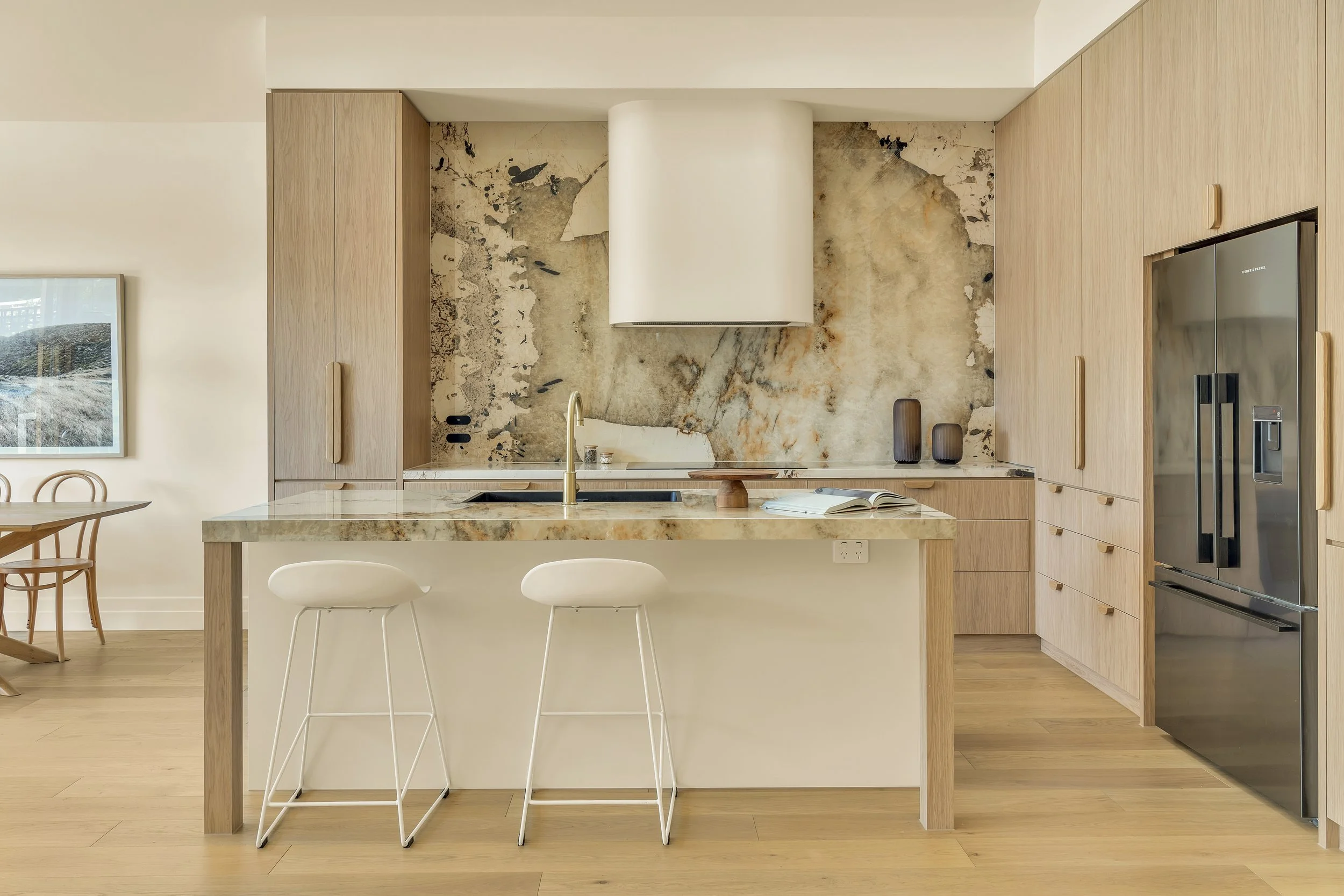 Modern kitchen with wooden cabinets, marble island, and two white bar stools. Features a marble backsplash, a gold faucet, and a stainless steel refrigerator.  Space designed by Aimee Bliss of Studio Hue in Gippsland, Victoria.