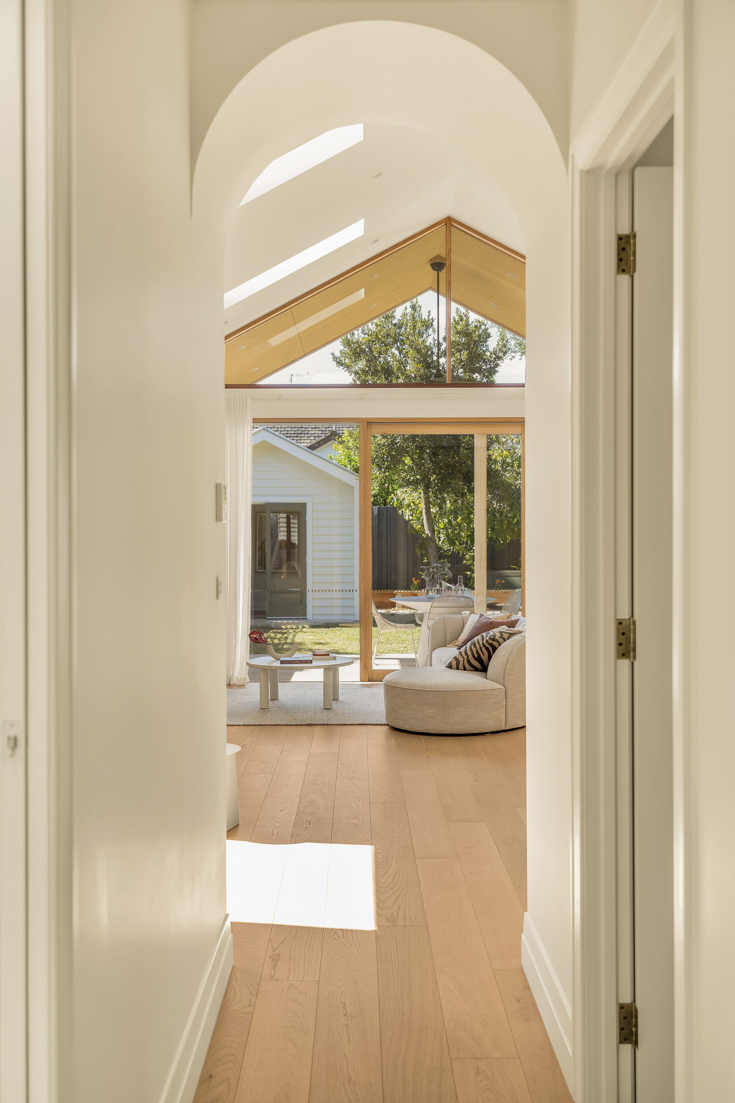 View through a doorway into a living room with large windows and a high, angled ceiling with skylights. The room has light-colored walls, wooden floors, a beige curved sofa with pillows, a small white coffee table with books and a decorative item, a 