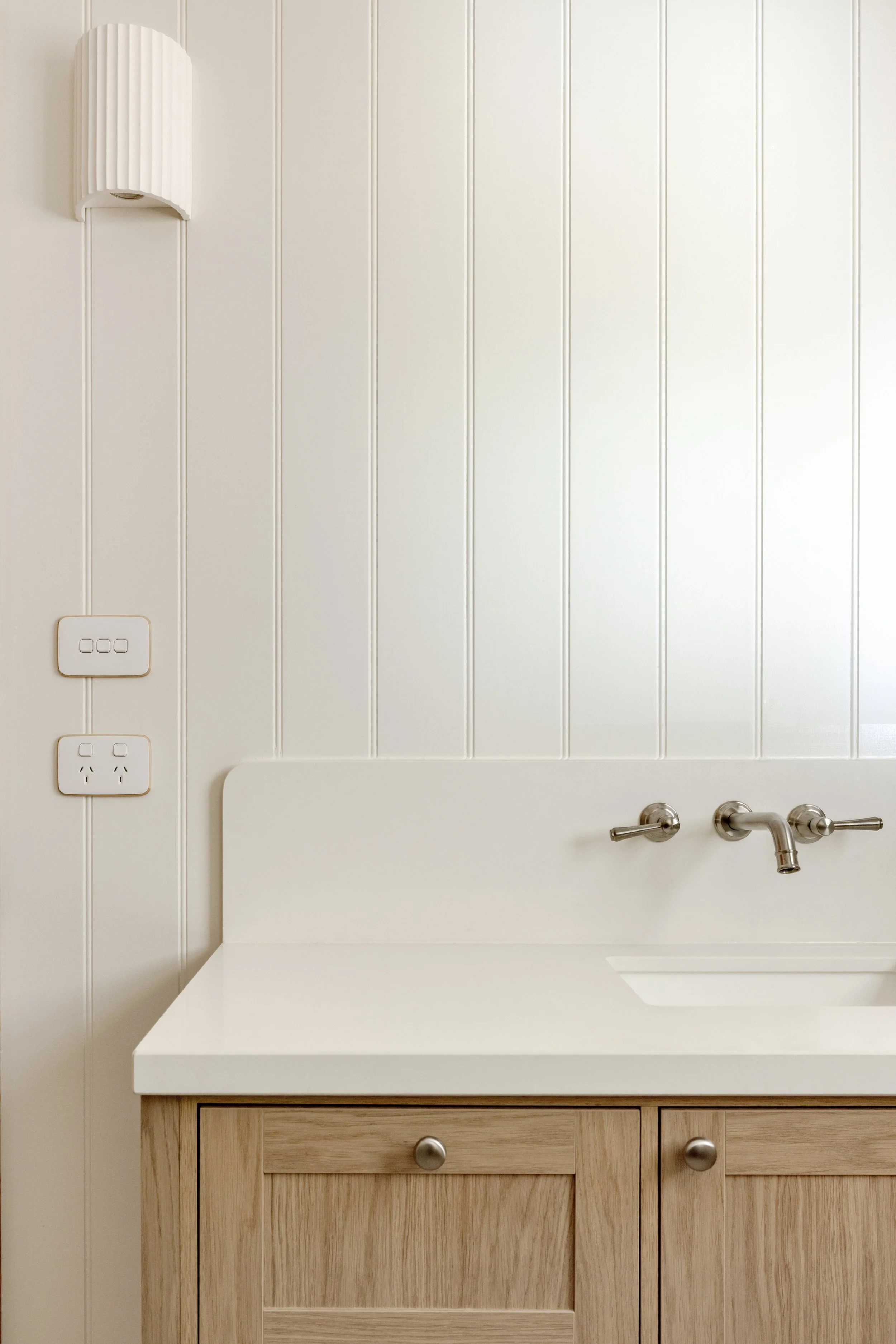Bathroom sink with a wooden vanity, a white countertop, a stainless steel faucet, white paneled wall, electrical outlets, and a white wall-mounted light fixture.