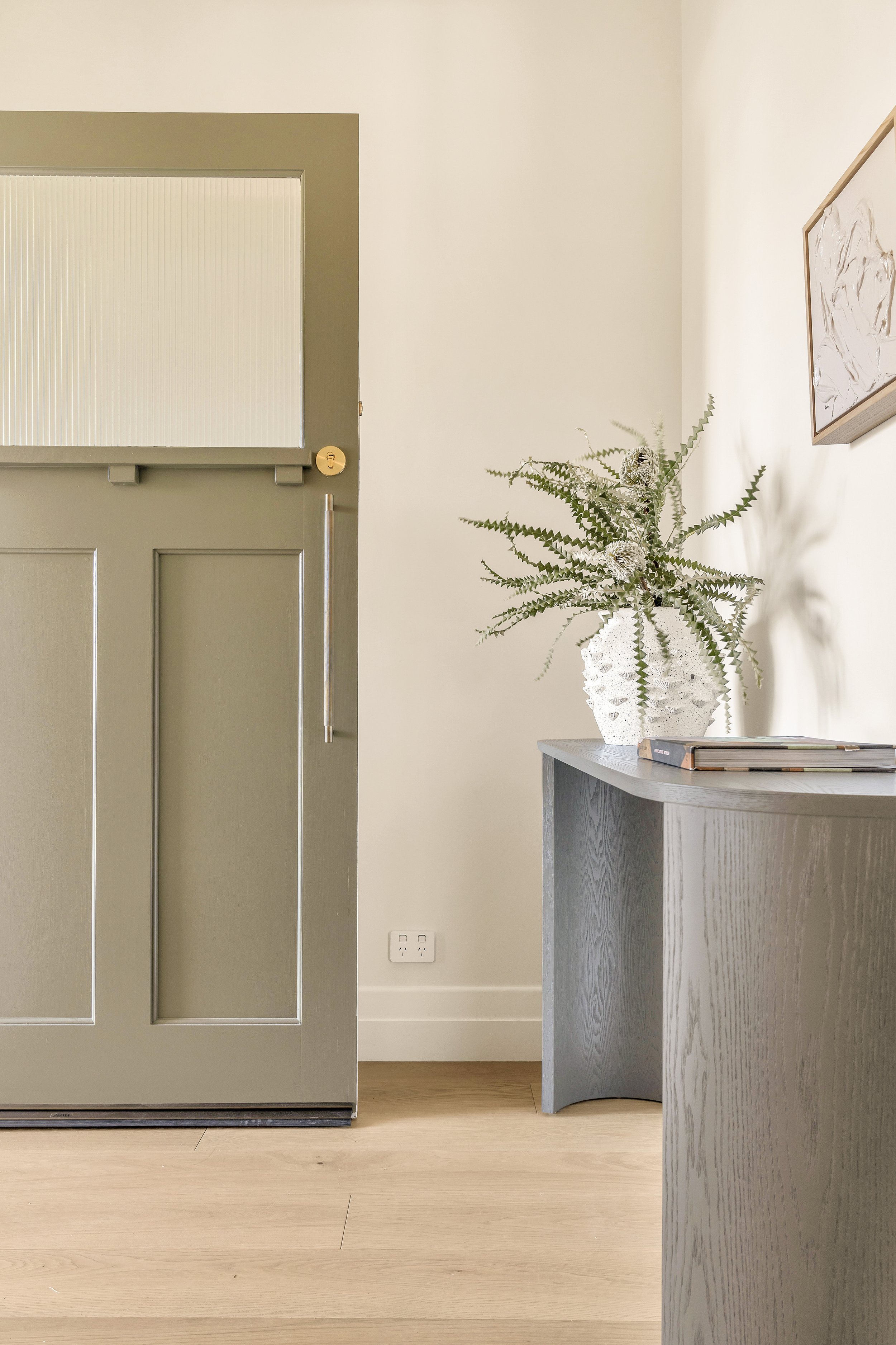 Interior scene with a green front door, a white textured vase holding a plant with long serrated leaves, part of a gray side table, and a framed abstract art piece on a white wall.