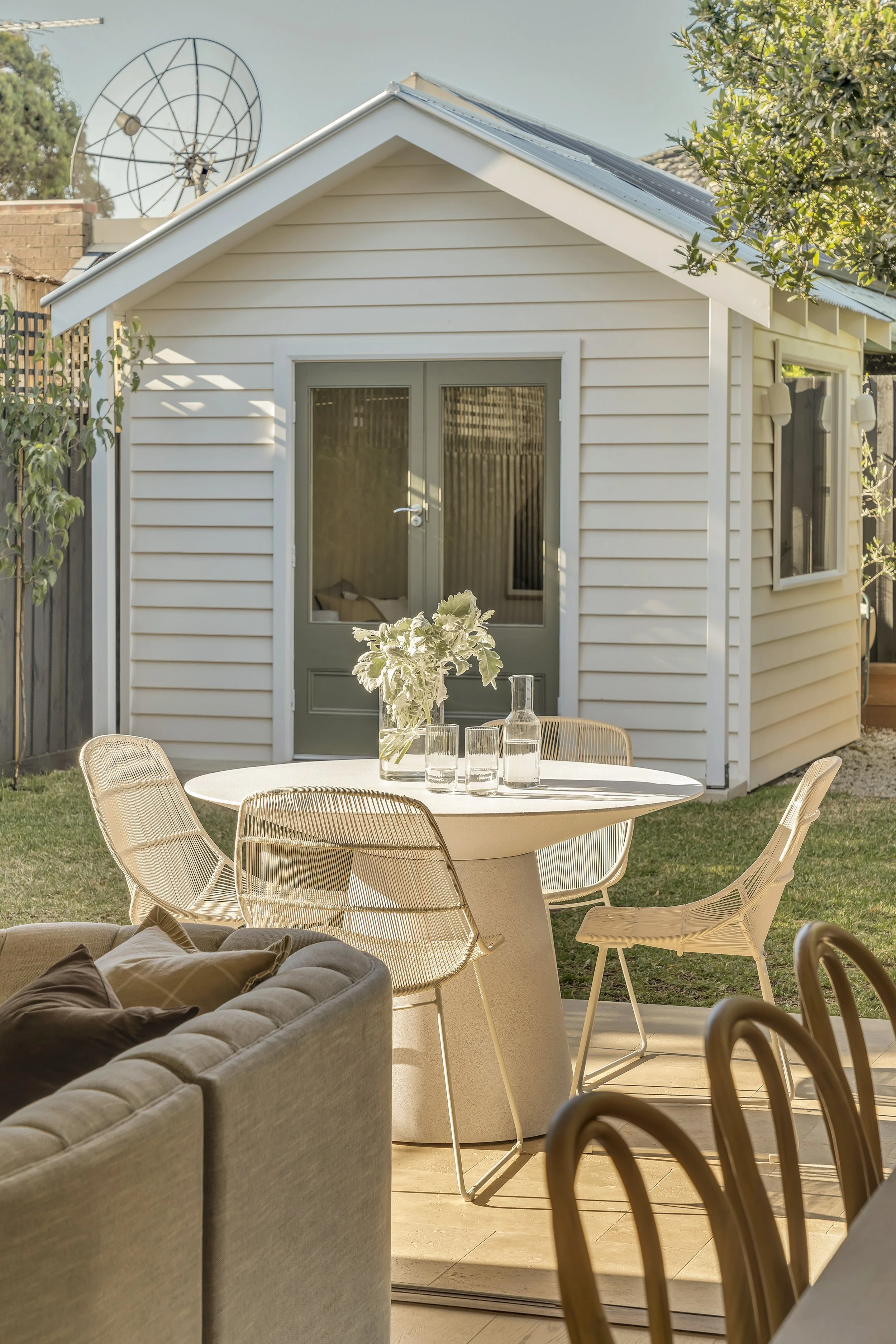 View of an outdoor patio area with a round white table and beige wicker chairs, a beige cushioned sofa with pillows, and a small white house with vinyl siding, glass doors, and windows in the background, surrounded by greenery.