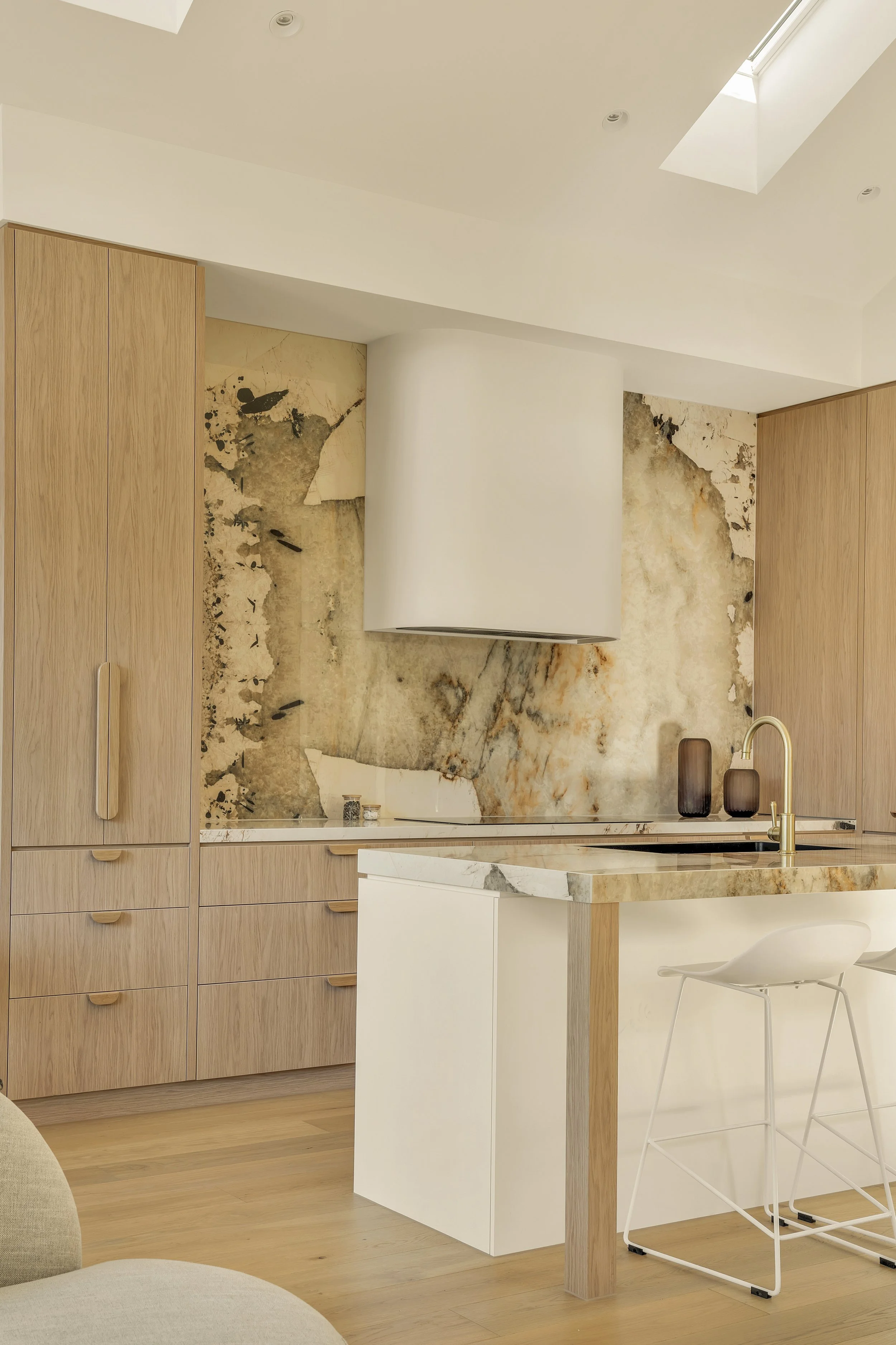 Modern kitchen with natural wood cabinets, a white marble island, and a marble backsplash, with barstools and decorative vases.
