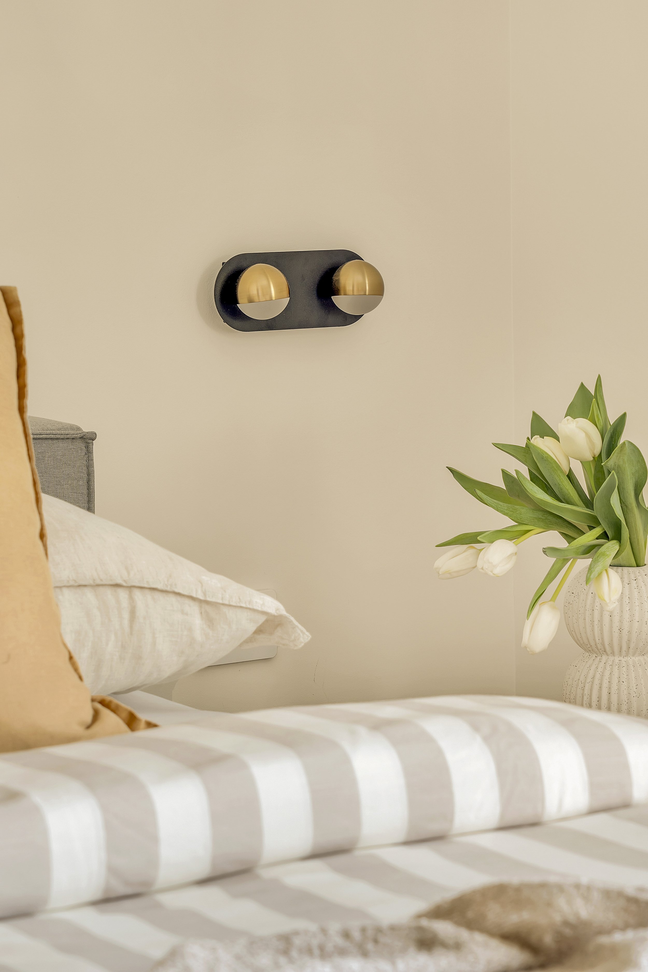Close-up of a bedroom with a beige wall, a black and gold wall light fixture, a gray headboard, a white pillow, and a vase with white tulips on a bed with a beige and white striped blanket.