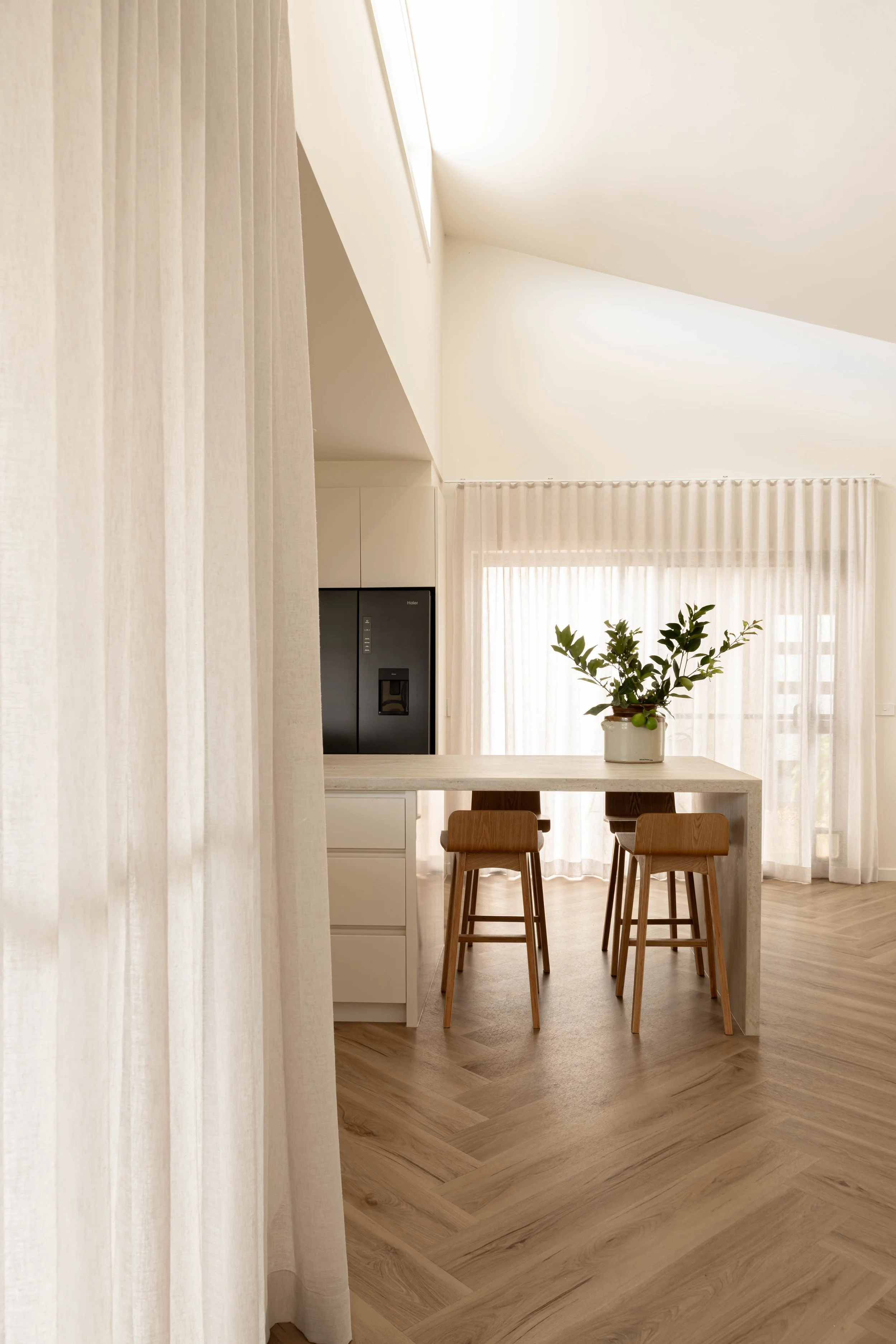 A minimalist kitchen with a dining area, featuring a white table and wooden chairs. The space is bright with sunlight filtering through sheer curtains, a potted plant on the table, and beige wooden flooring.