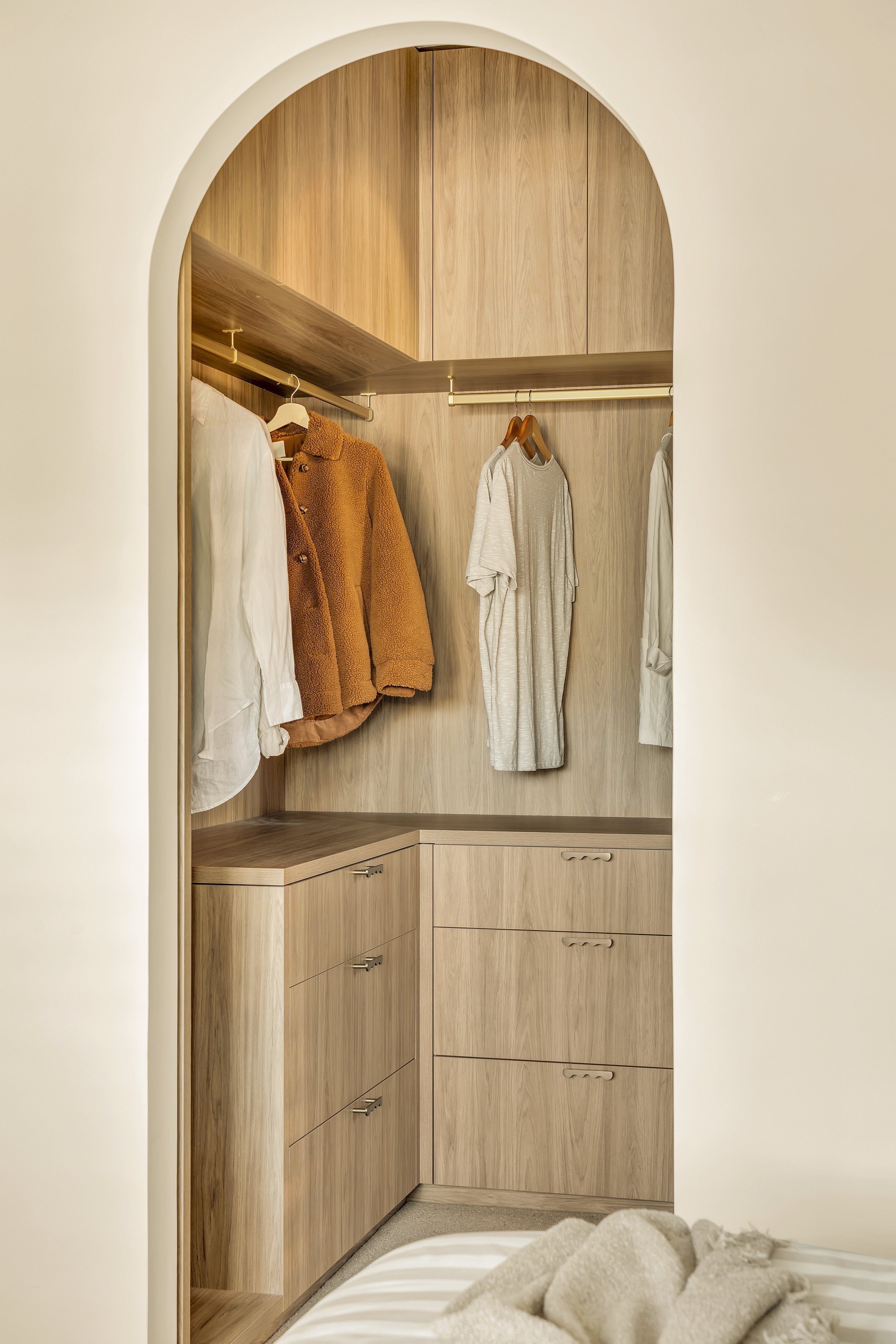 Inside a wooden wardrobe with clothes hanging on a rod, including a white shirt, orange coat, and gray dress, with drawers at the bottom and an arched entryway.