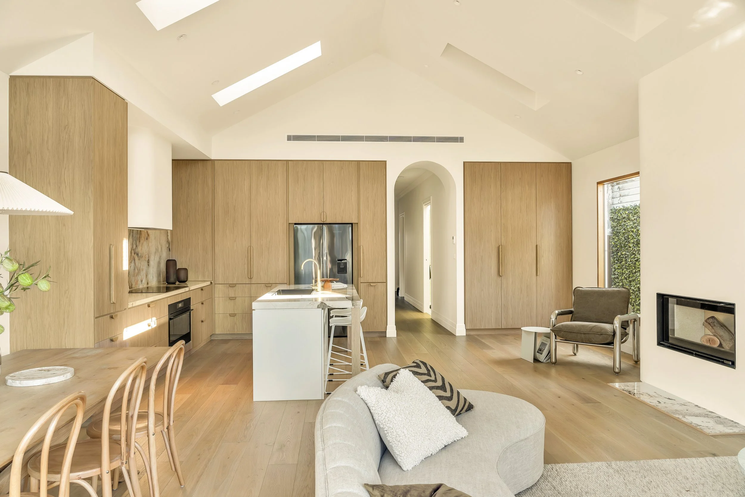 Open-concept living space with kitchen, dining, and seating area. Wooden cabinets, kitchen island, and skylights with a cozy seating area featuring a beige sofa and armchair. Large window with view of greenery.