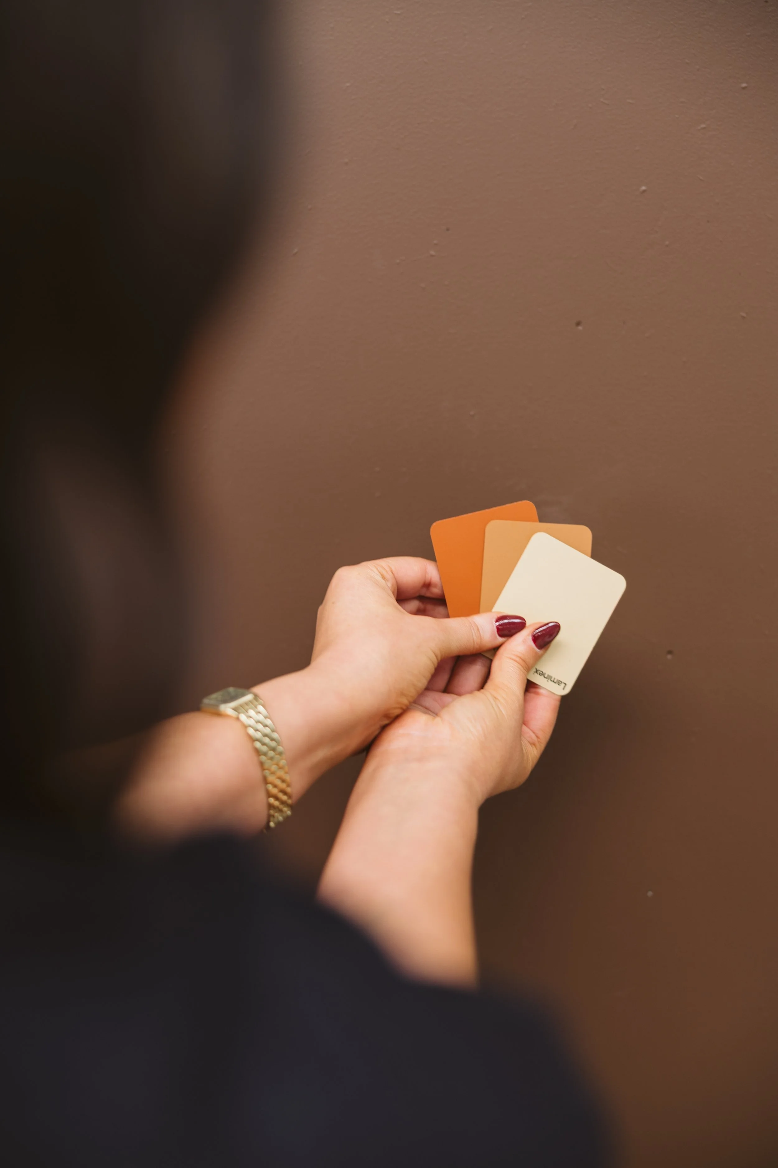 A person's hand holding three color swatch cards against a brown wall. The person is wearing a gold bracelet and has red nail polish.
