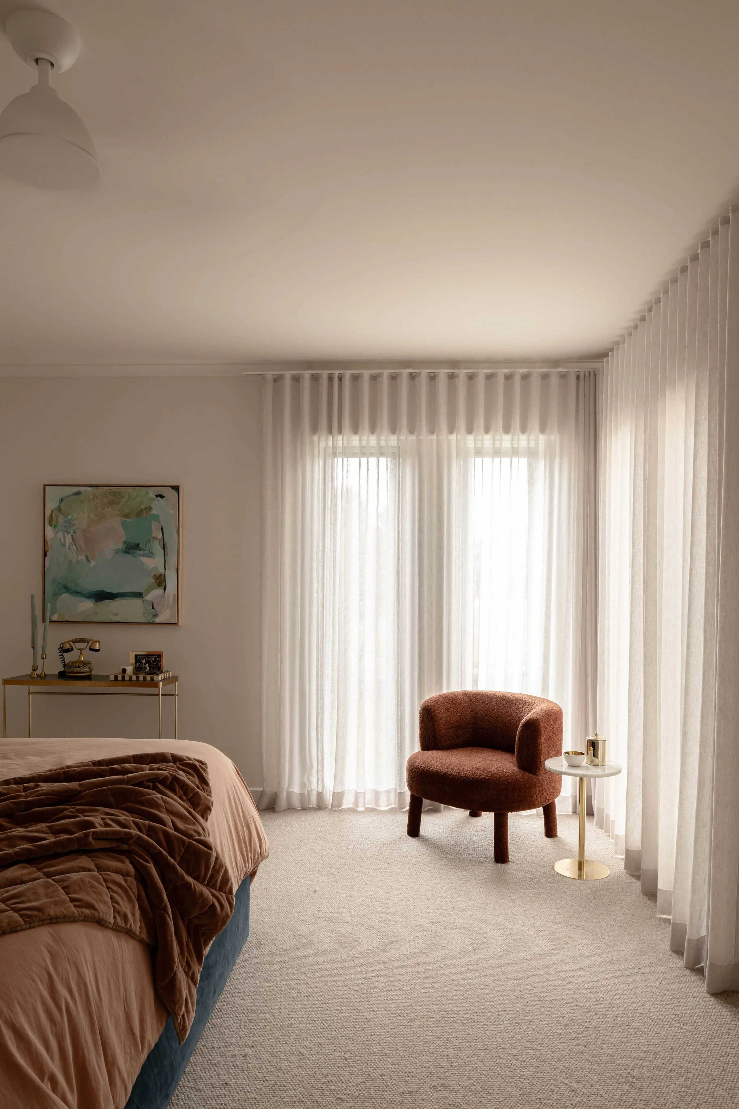 A bedroom with beige curtains, a brown armchair, a light-colored carpet, a bed with a brown cover, a side table with a decorative item, and wall art.