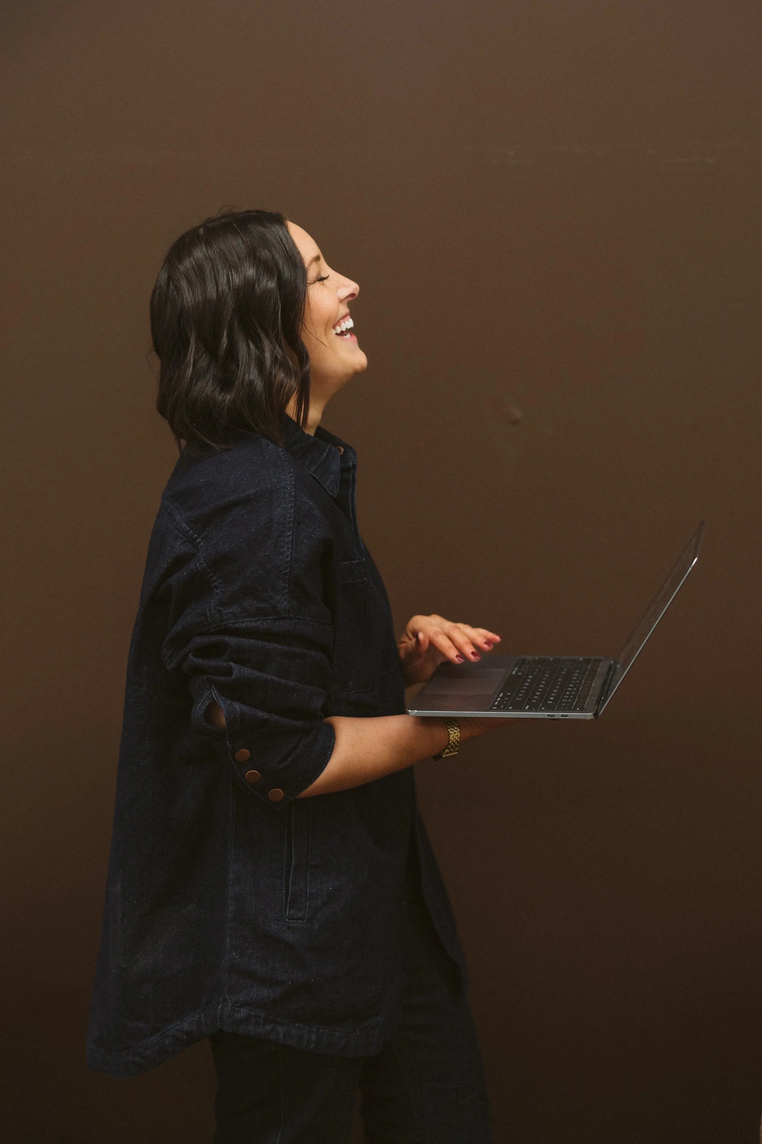 Photo of Aimee Bliss of Studio Hue in Gippsland, Victoria.  She has dark wavy hair and is smiling, holding a laptop, standing against a brown background.