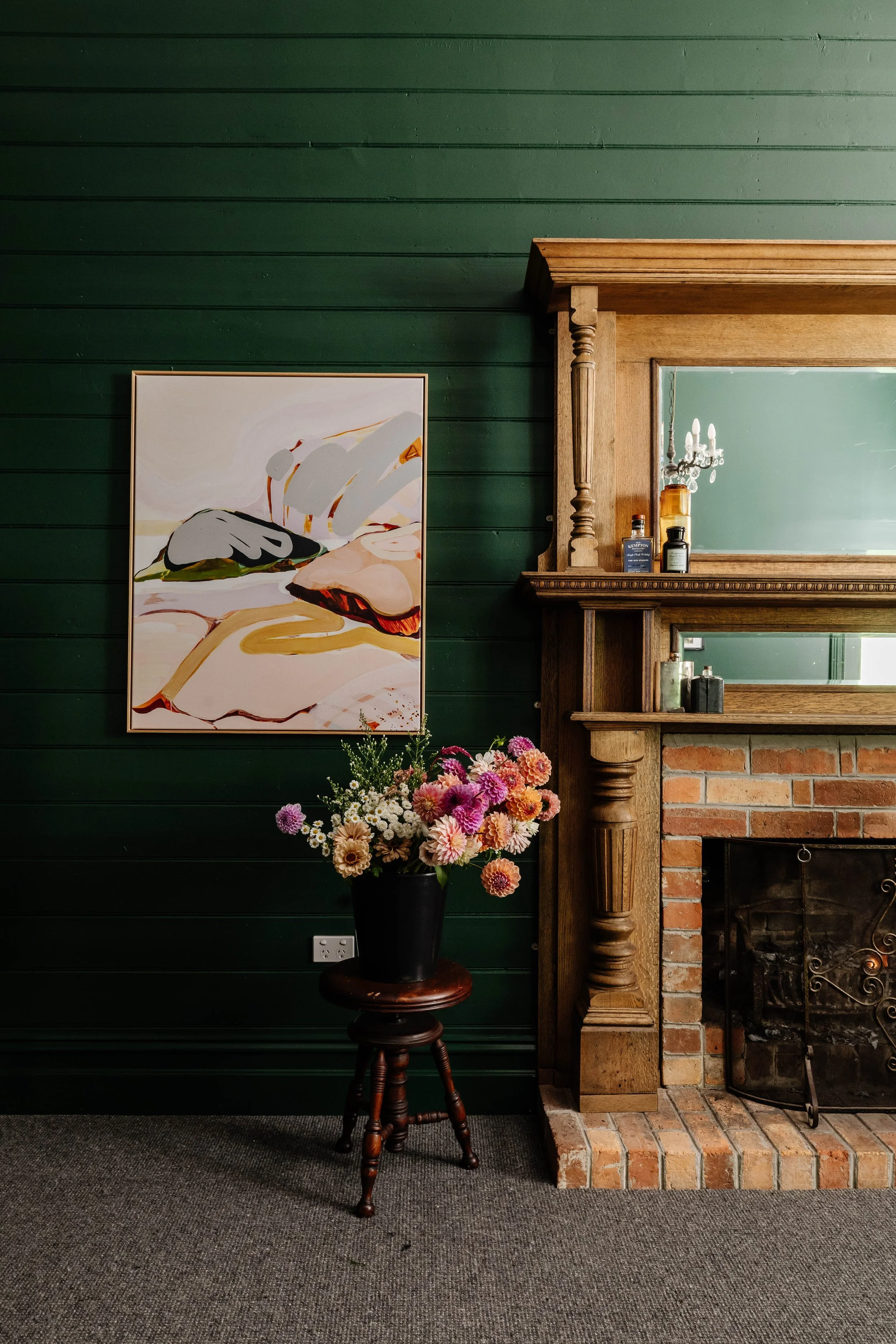 A room with dark green wooden paneled wall, a colorful abstract painting on the left, a wooden fireplace with mirror on the right, a black vase with pink and white flowers on a wooden stool in front of the painting, and a dark carpeted floor.
