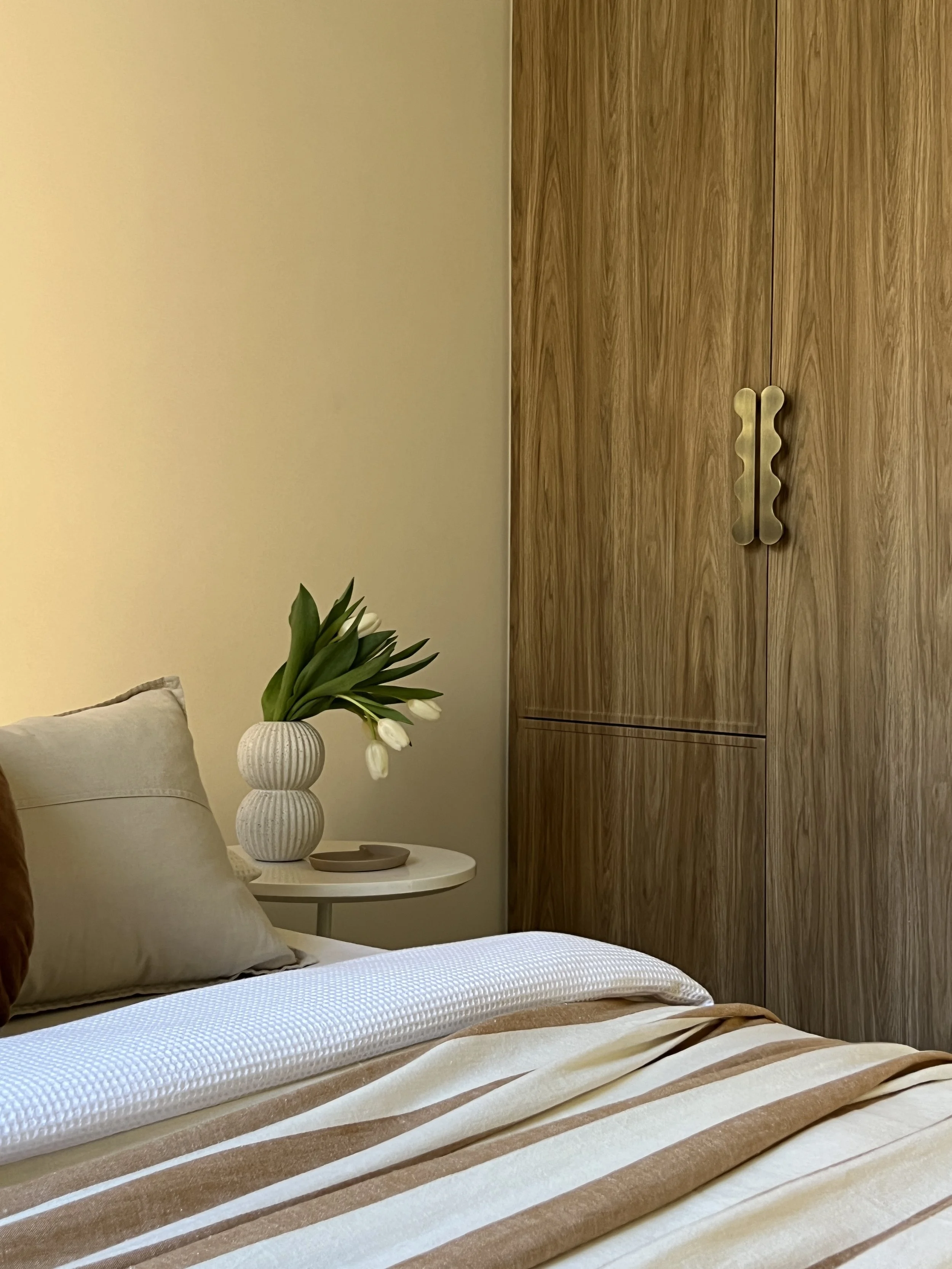 A bedroom corner featuring a bed with beige and striped bedding, a white side table with a vase of white tulips, and a wooden wardrobe with metallic handles.