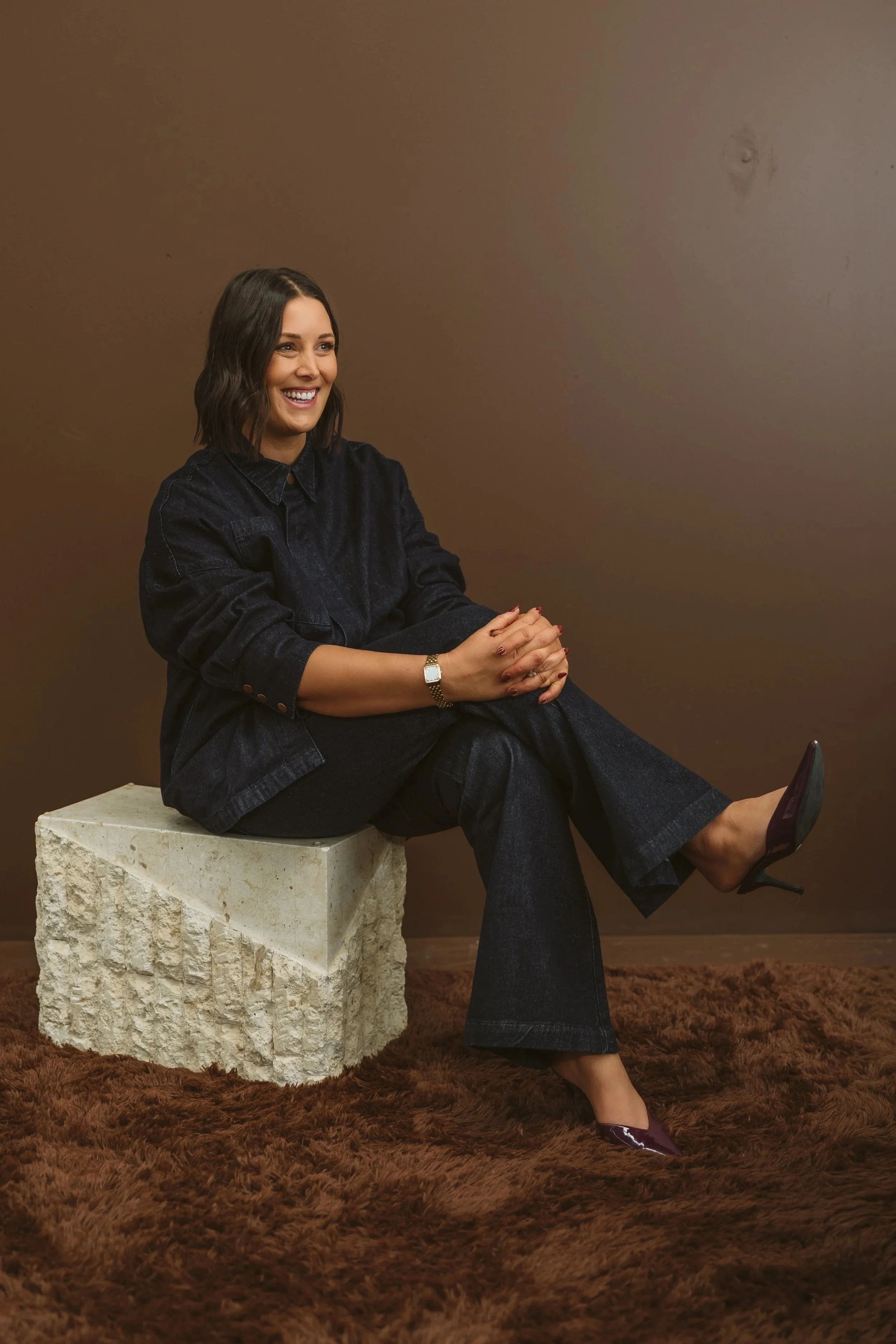 Aimee Bliss, from Studio Hue Interior Design.   She has shoulder-length dark hair and iseated on a stone block, smiling, wearing a dark denim shirt, dark pants, and purple high heels, in a room with a brown wall, brown shag carpet, and a small table.