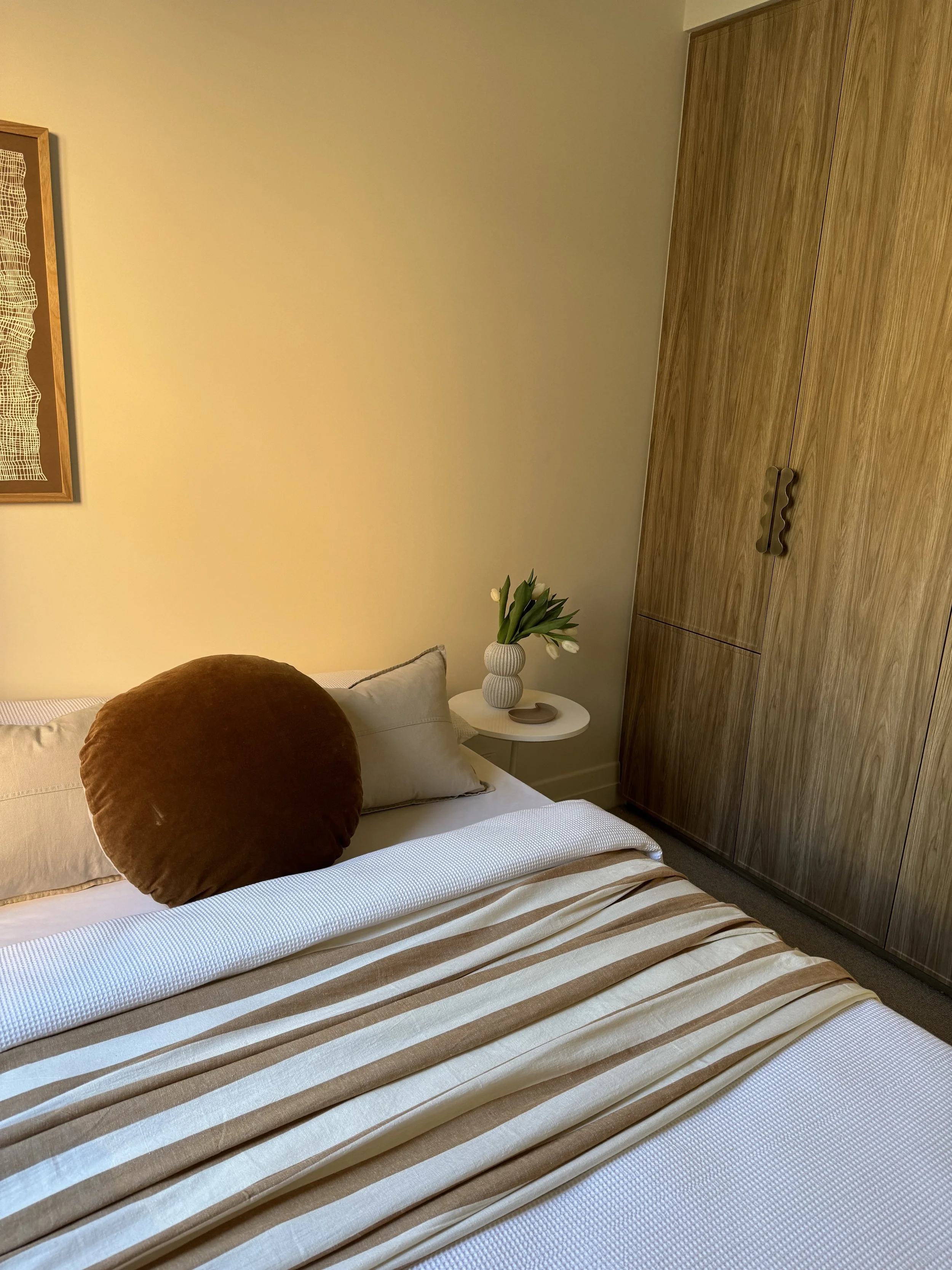 A neatly made bed with beige and brown striped bedspread, two beige pillows, and one round brown pillow, beside a small round white bedside table with a vase of white tulips. A large wooden wardrobe is in the corner, and a framed artwork hangs on the cream-colored wall.