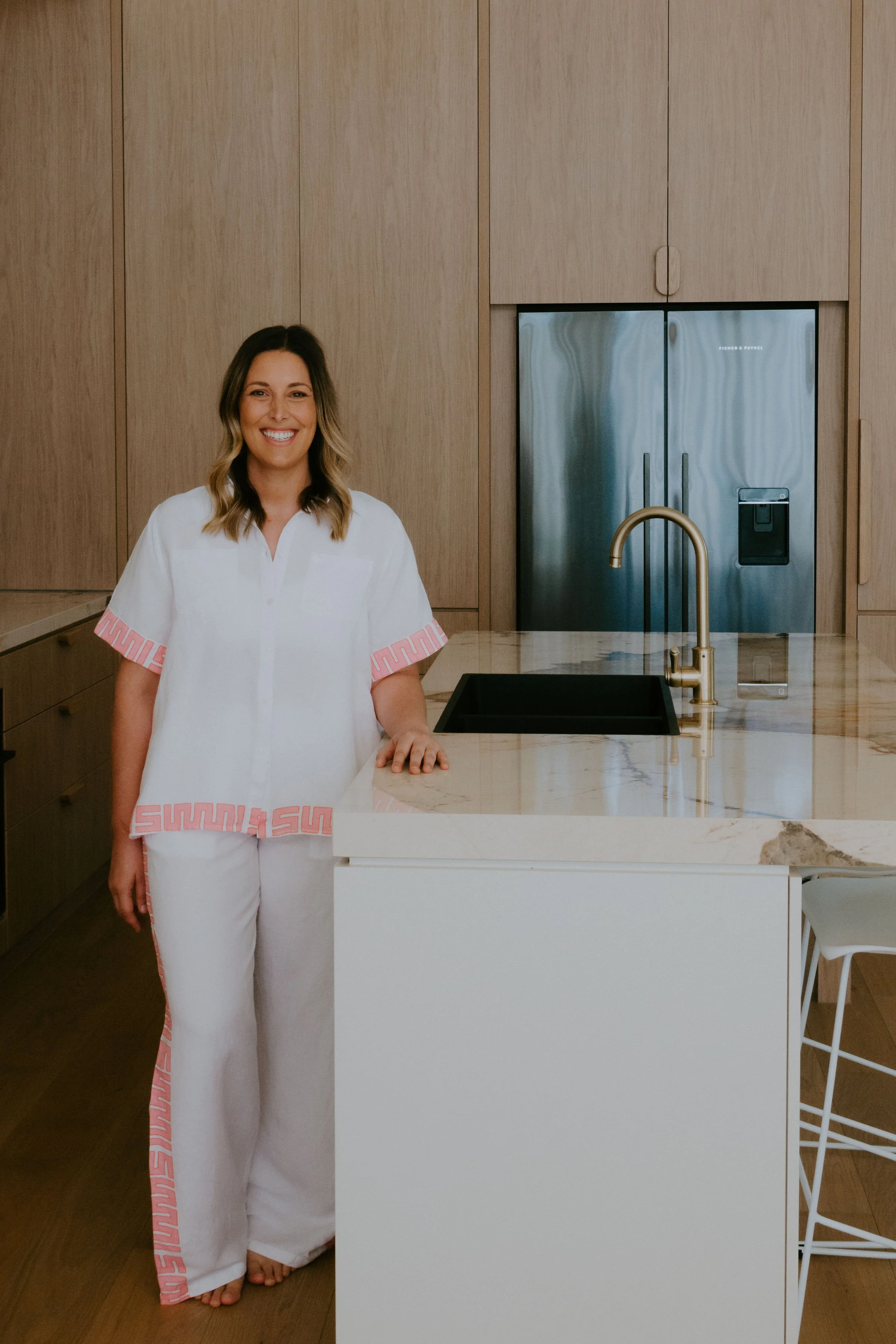 A woman with shoulder-length brown hair, smiling, stands in a modern kitchen next to a marble island with a black sink and gold faucet, wooden cabinets, and a stainless steel refrigerator.