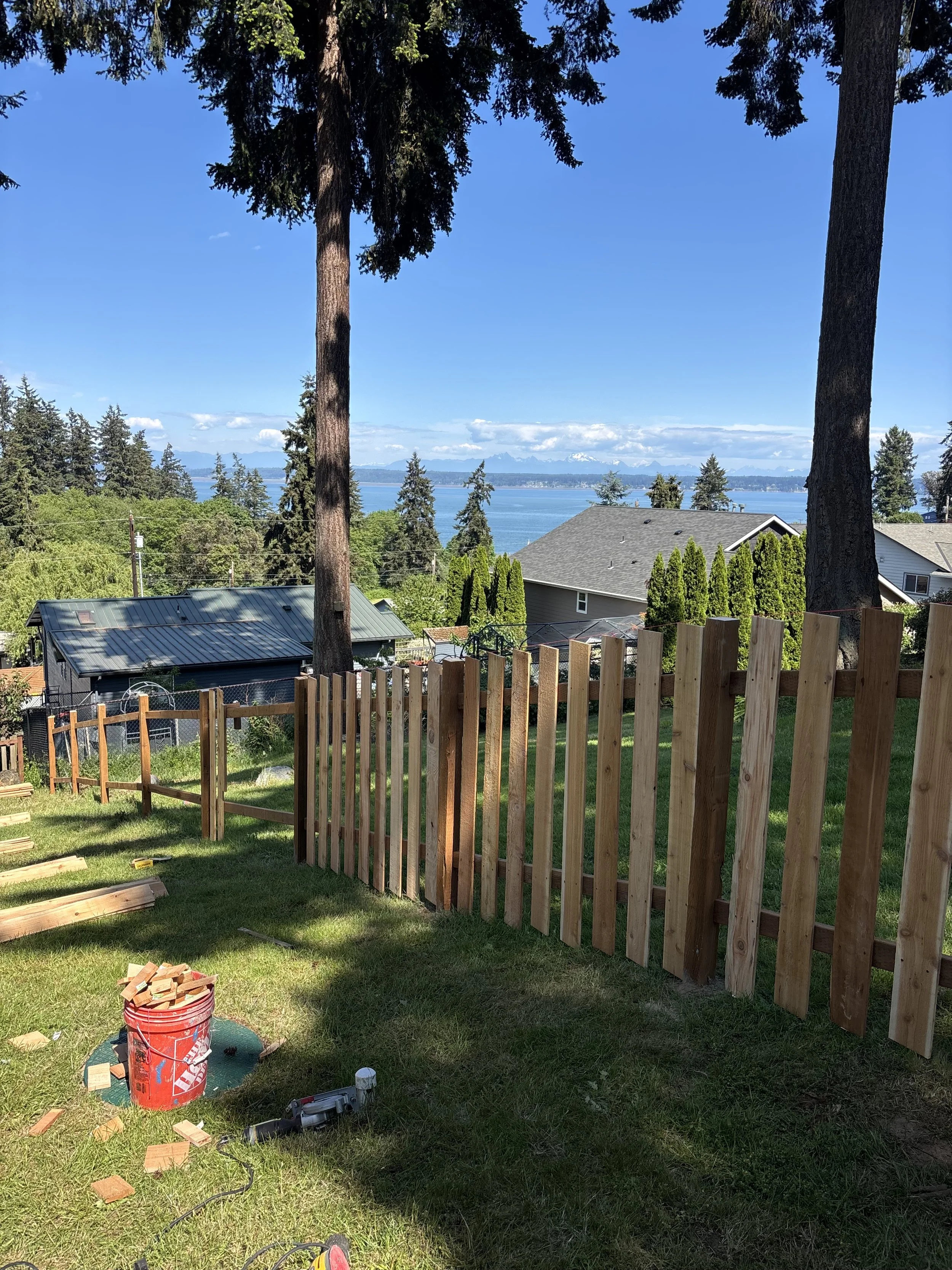 A backyard has a wooden fence, construction tools, and an area under construction with a view of trees and water in the distance under a blue sky with clouds.