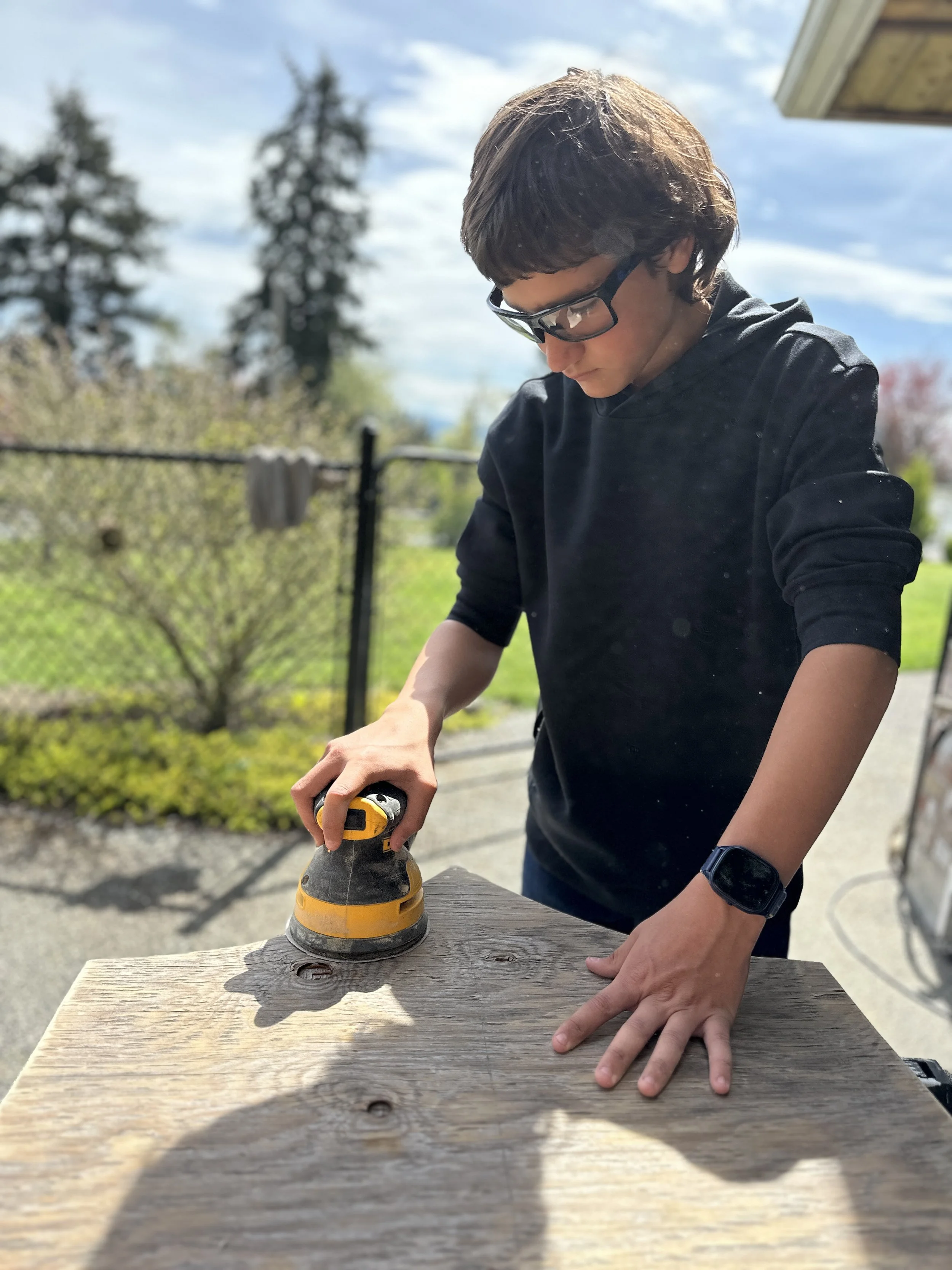 A person wearing glasses and a black hoodie is sanding a wooden surface outdoors. They are using a power sander with their right hand while their left hand holds the wood steady. The background shows trees and a cloudy sky.