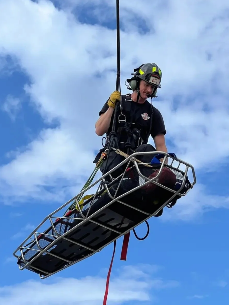 A firefighter in protective gear riding in a hanging rescue basket during a training exercise or emergency response, against a backdrop of blue sky and clouds.