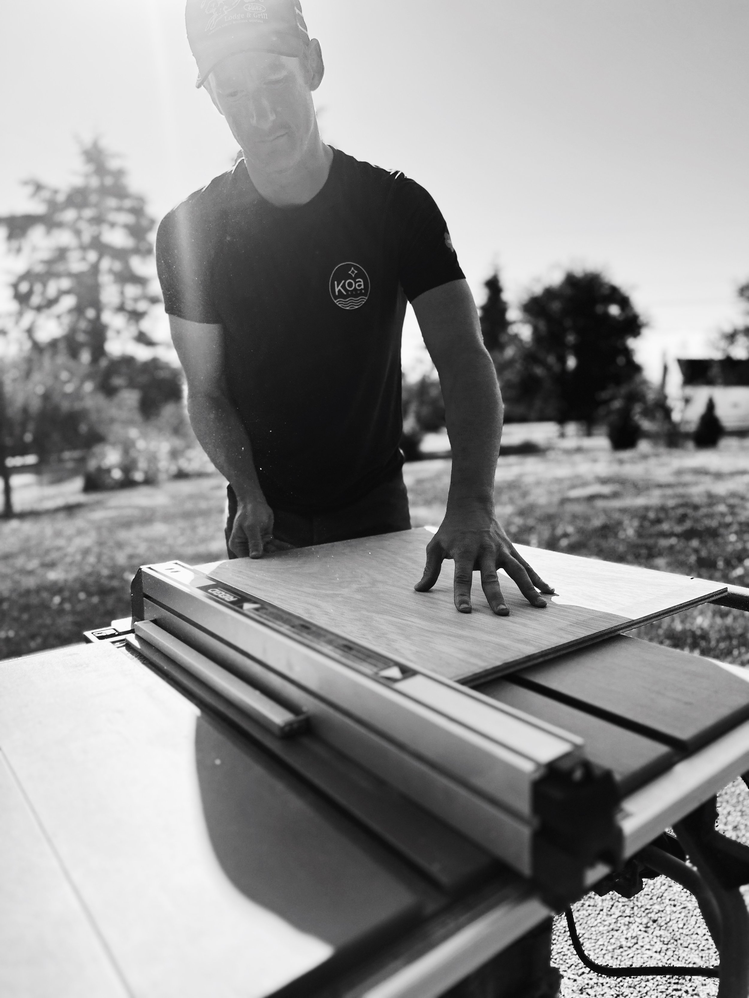 A man standing outside in front of a table, using a hand saw to cut a piece of wood.