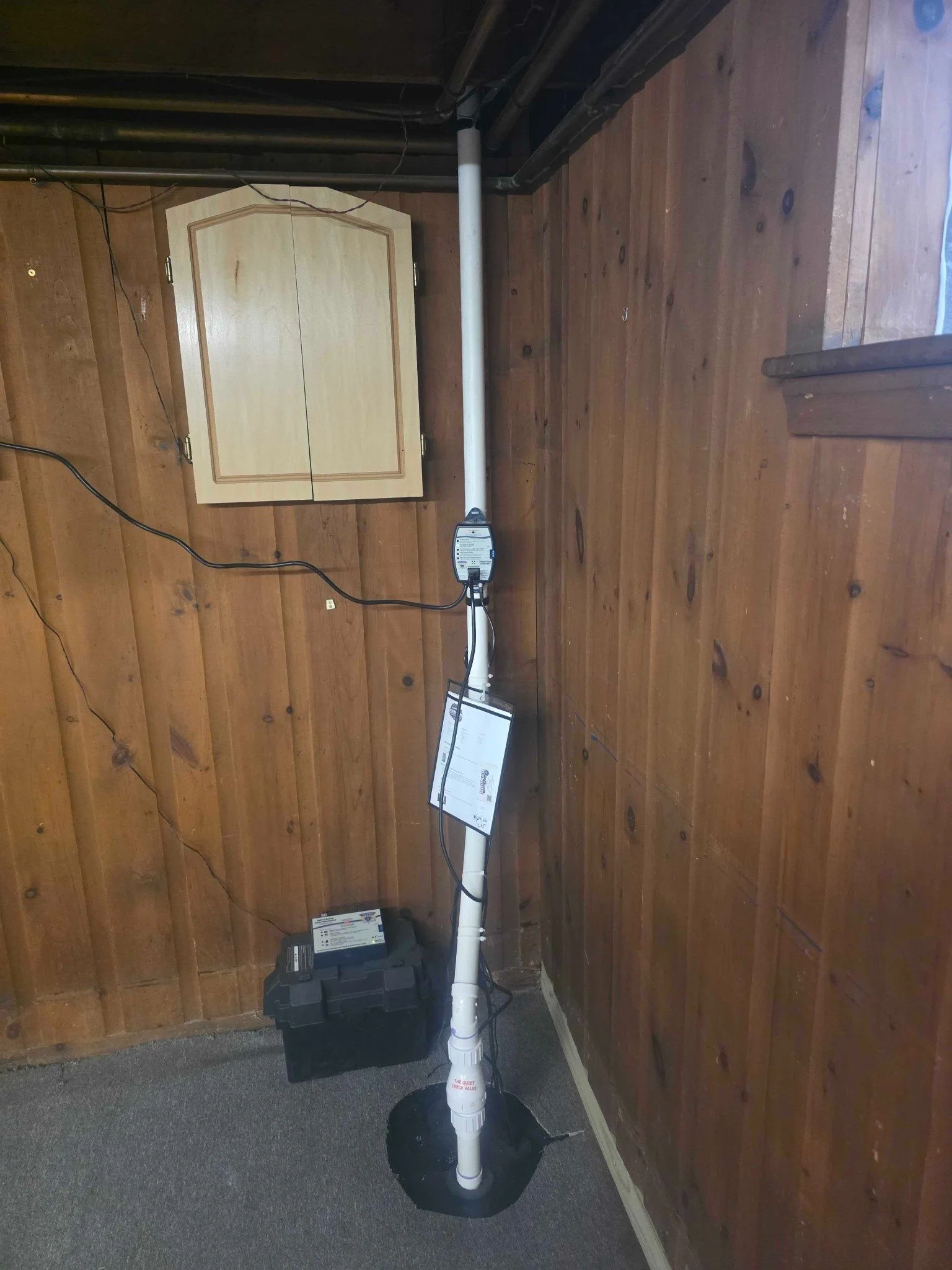 Completed sump pump discharge piping and restored wood-panel wall in the finished basement.