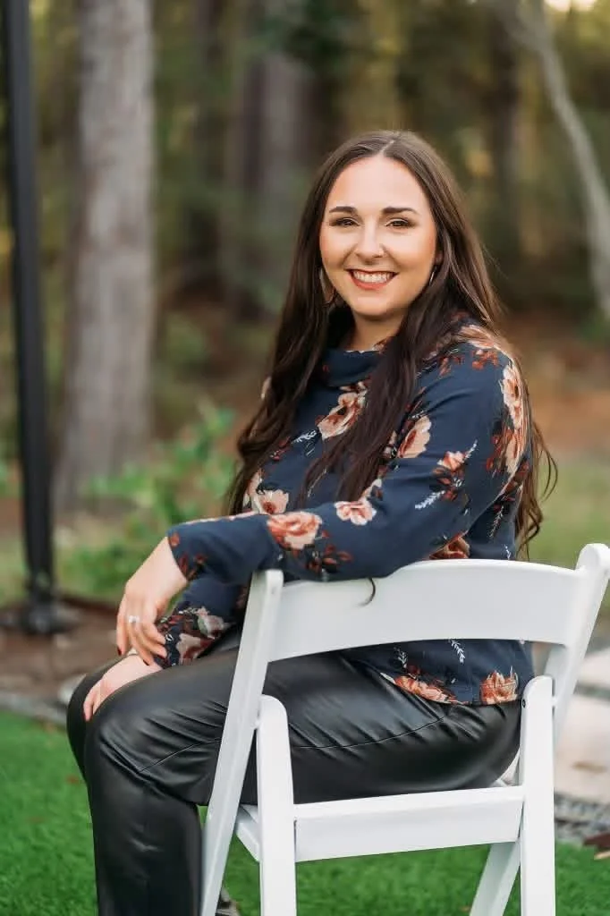 A woman with long dark hair smiling and wearing a floral blouse is sitting on a white chair outdoors, with trees and greenery in the background.