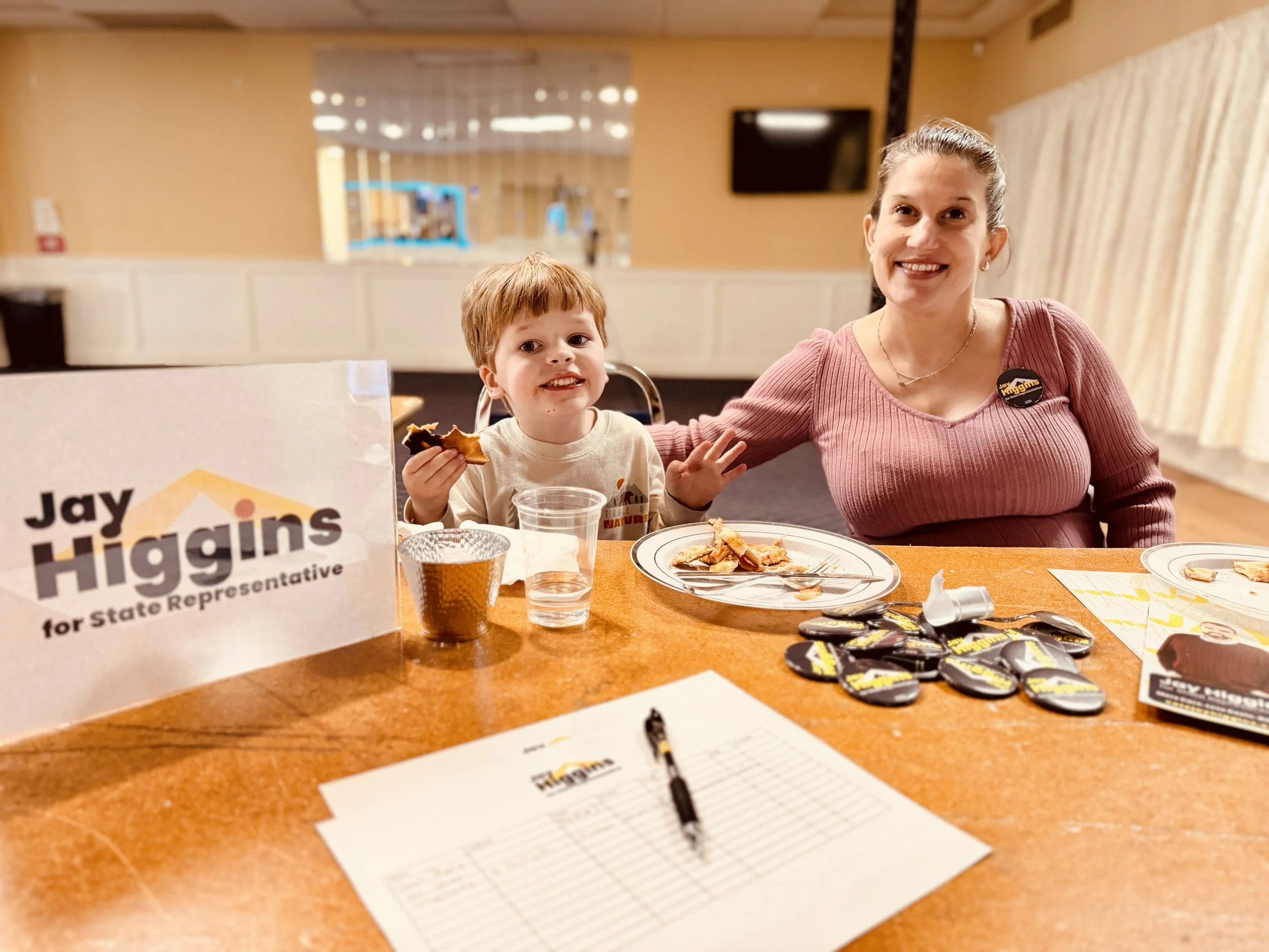 Two volunteers sit at a welcome desk. One is a child, smiling with a half eaten piece of pizza. The other is an adult woman wearing a pink dress and smiling with her arm around the child. On the table are Jay Higgins for State Rep materials.