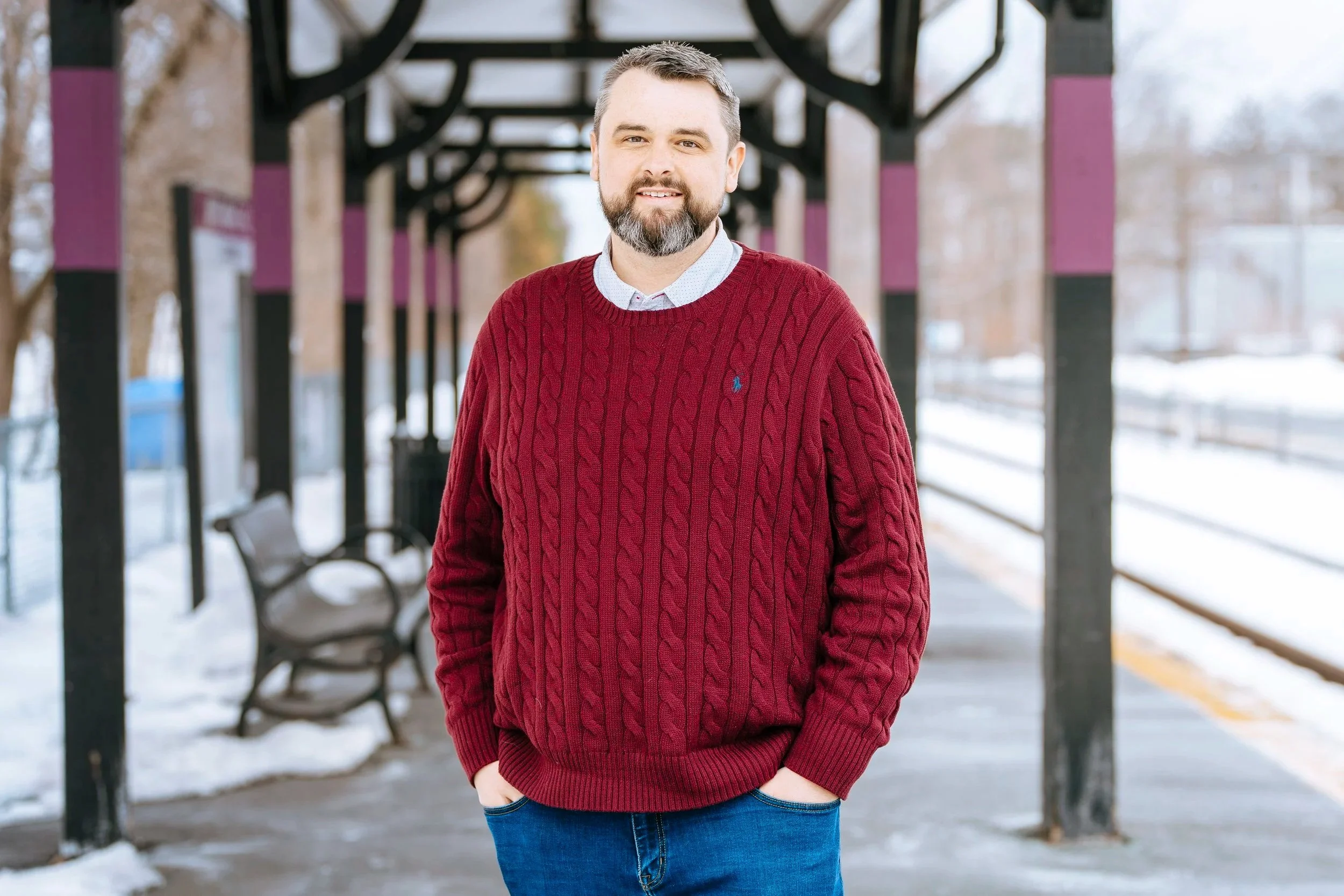 A portrait of Jay Higgins standing at the Wyoming Commuter Rail station. Jay is wearing a red sweater and jeans.
