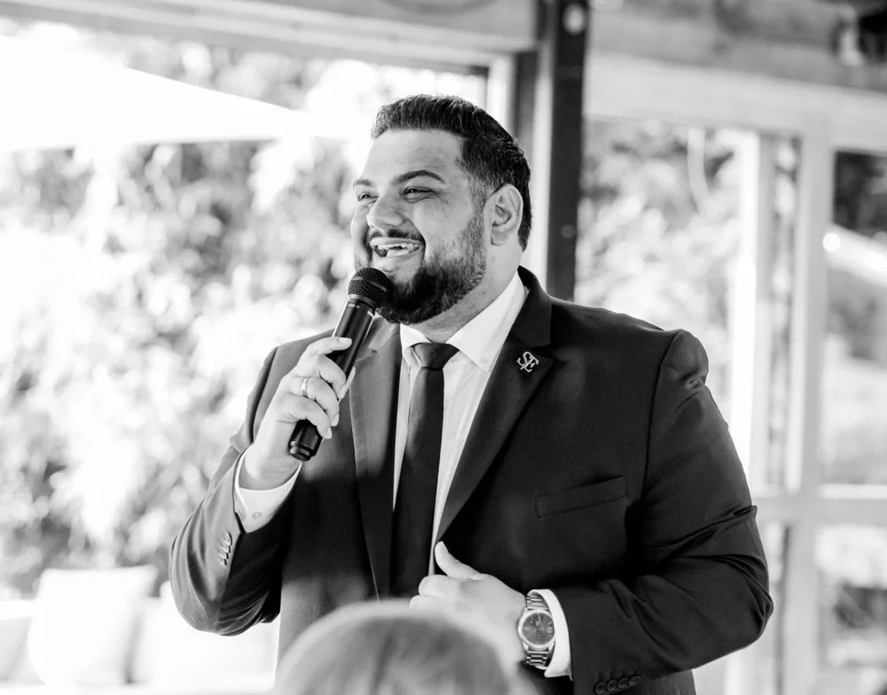 Der freie Trauredner Pedi in einem Anzug mit Krawatte hält ein Mikrofon und spricht bei einer Veranstaltung. Er trägt eine Uhr und einen Anstecker am Revers.