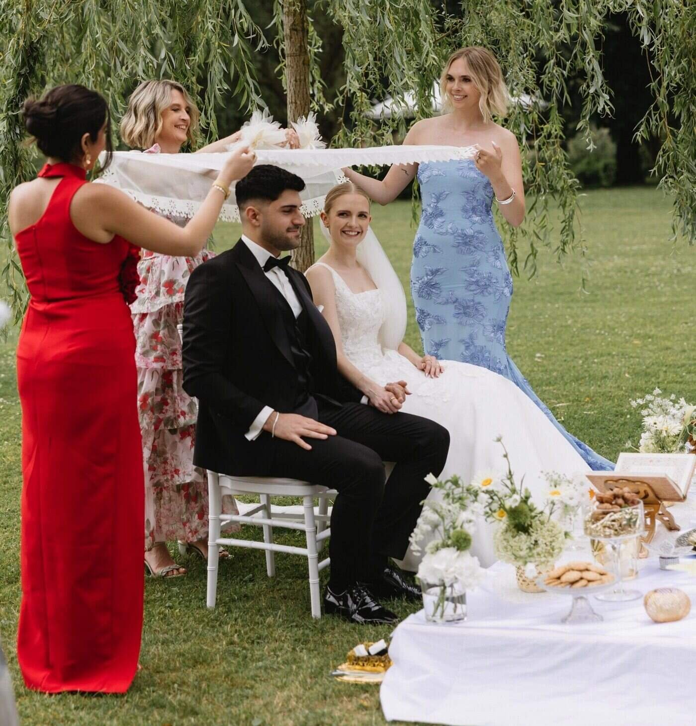 Eine Hochzeitszeremonie im Freien mit dem freien Trauredner Pedi, mit einem Brautpaar sitzend und Freunden, die einen Schleier über sie halten.