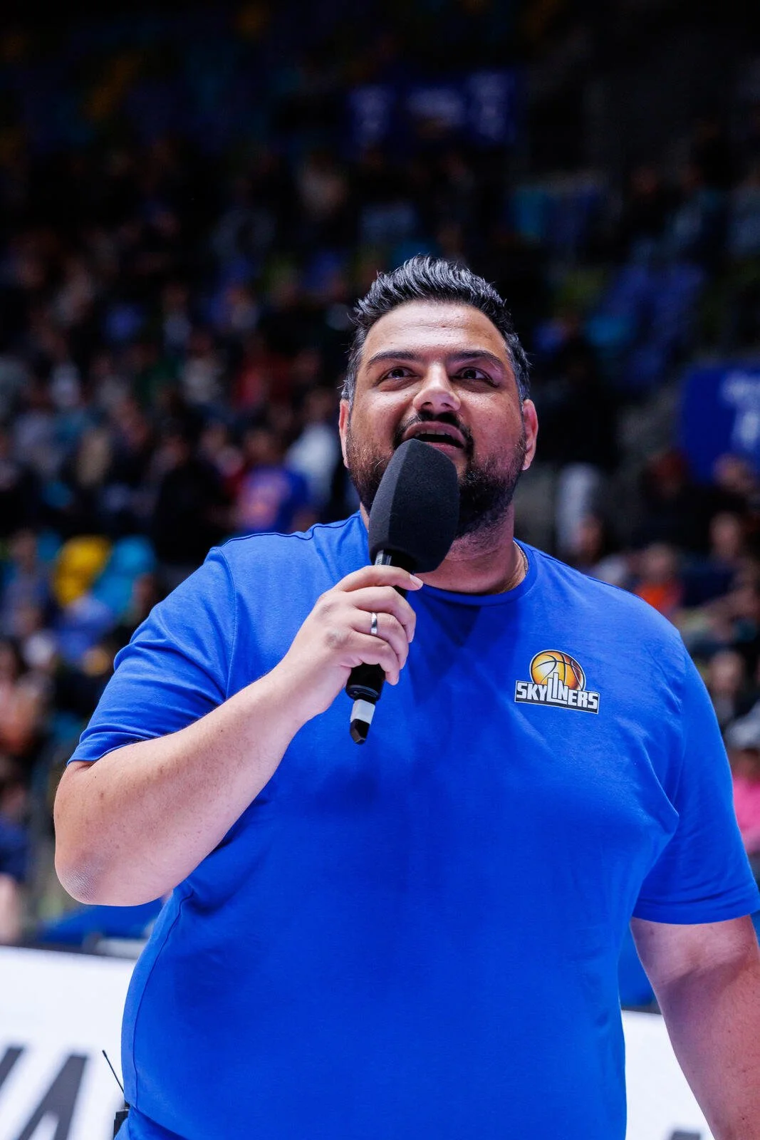 Ein Mann mit dunklem Haar und Bart, der in einem blauen Skyliners-T-Shirt einen schwarzen Mikrofon hält, steht vor einem großen Publikum in einer Halle.