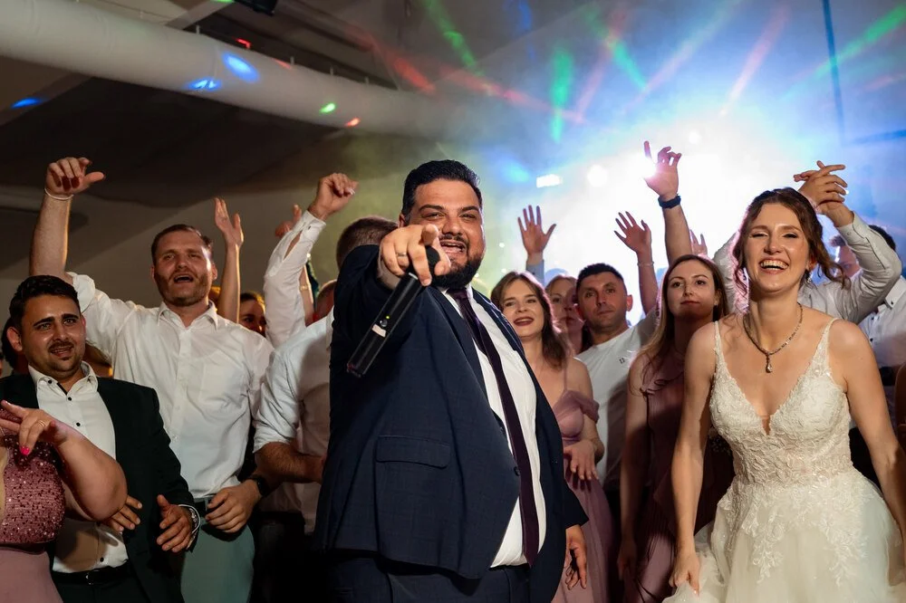 Menschen feiern auf einer Party, mit bunten Lichtern und fröhlicher Stimmung. Im Vordergrund ein Mann in dunklem Anzug, der in die Kamera lächelt und mit einem Mikrofon posiert. Neben ihm eine lachende Frau in einem weißen Kleid, wahrscheinlich Braut. Die Gruppe im Hintergrund hebt die Hände in die Luft und freut sich.