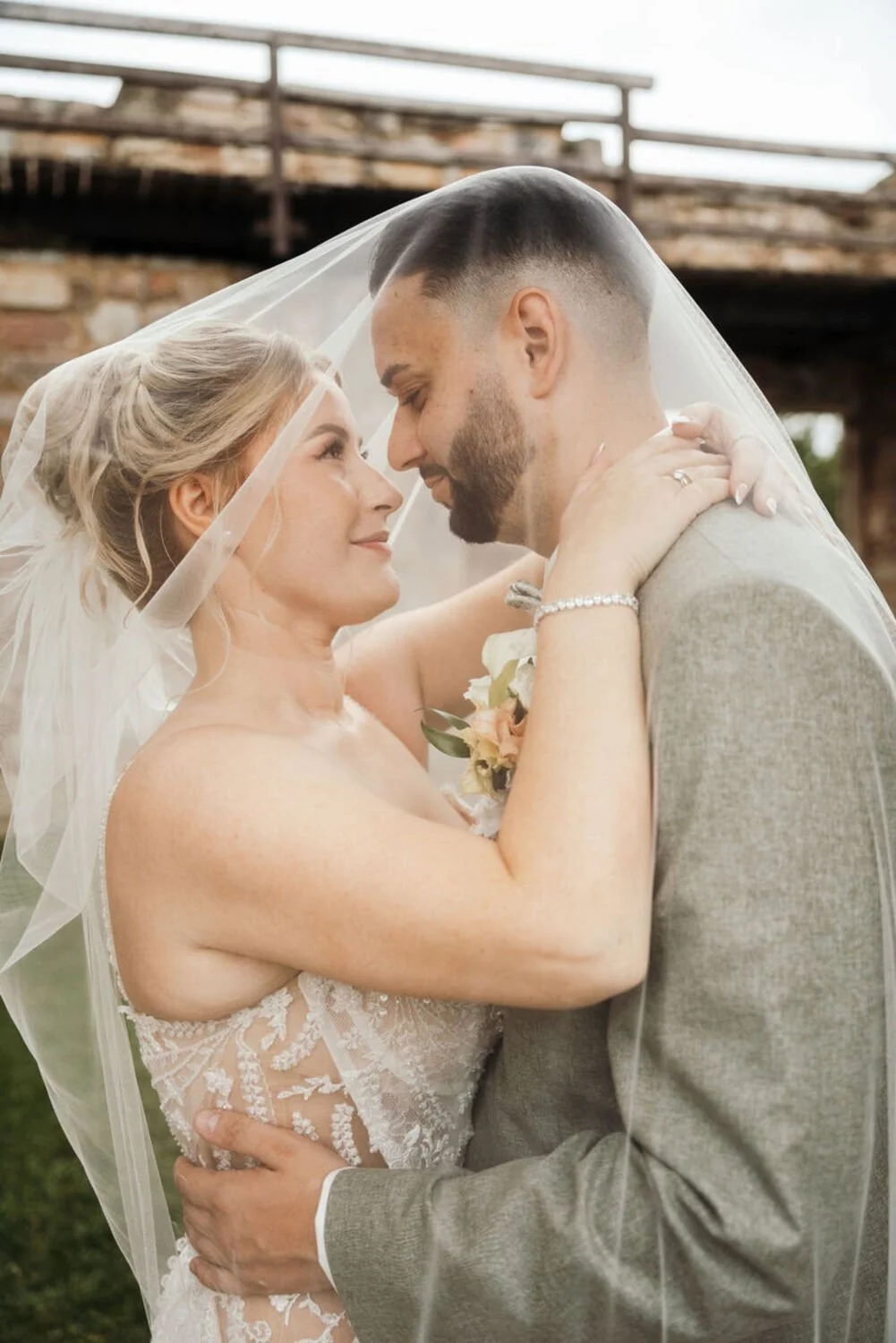 Ein Brautpaar umarmt sich liebevoll, sie trägt ein Brautkleid mit Spitze, er trägt einen grauen Anzug, beide blicken sich in die Augen unter einem Schleier bei ihrer Hochzeit.