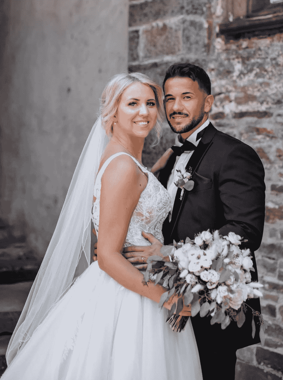 Ein heirates Paar in Hochzeitskleidung vor einer Steinmauer, die Braut hält einen Blumenstrauß.