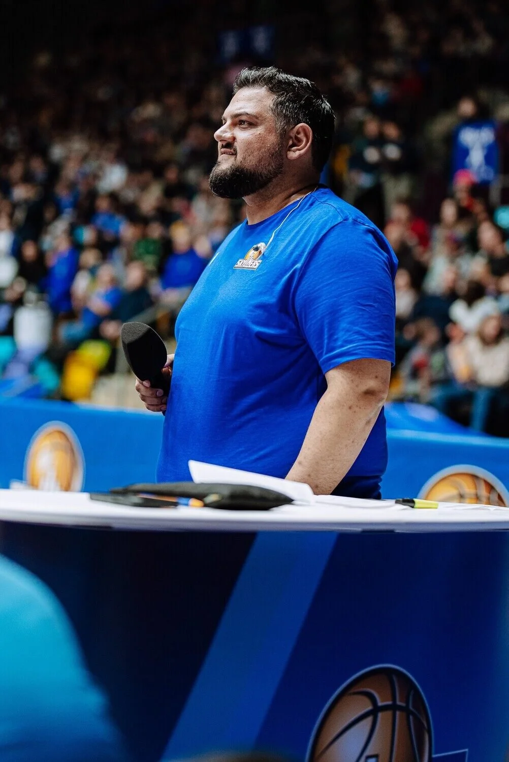 Der freie Trauredner Pedi in blauer Sportbekleidung mit Headset und Mikrofon, steht hinter einem Tisch bei einer Basketball-Veranstaltung, im Hintergrund ein Publikum.