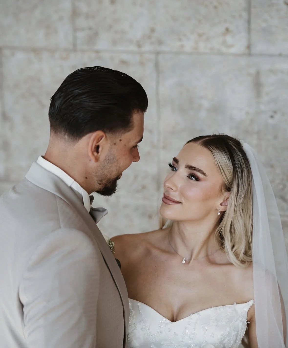 Ein Brautpaar bei ihrer Hochzeit, Blickkontakt, in eleganter Kleidung, vor einem grauen Steinmauer-Hintergrund.